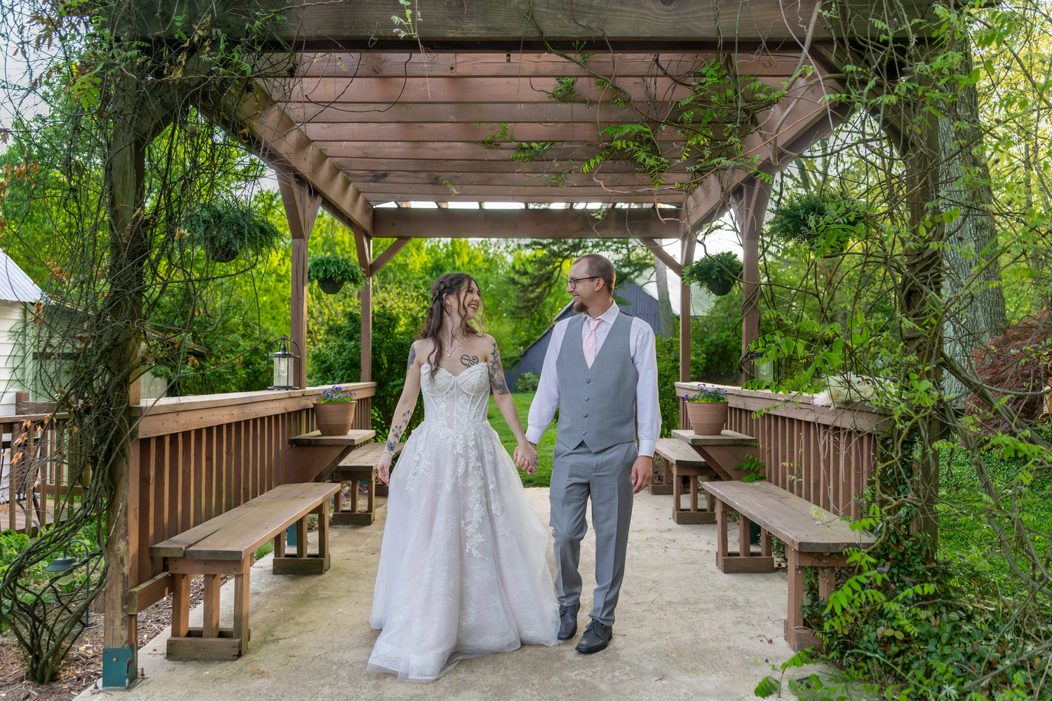Bride and groom hold hands walking under a wooden pergola surrounded by lush greenery at an outdoor wedding venue.