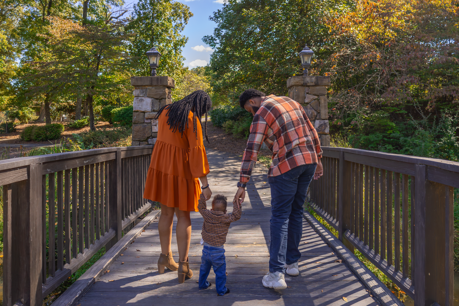 A family walks together holding hands on a wooden bridge surrounded by autumn foliage.