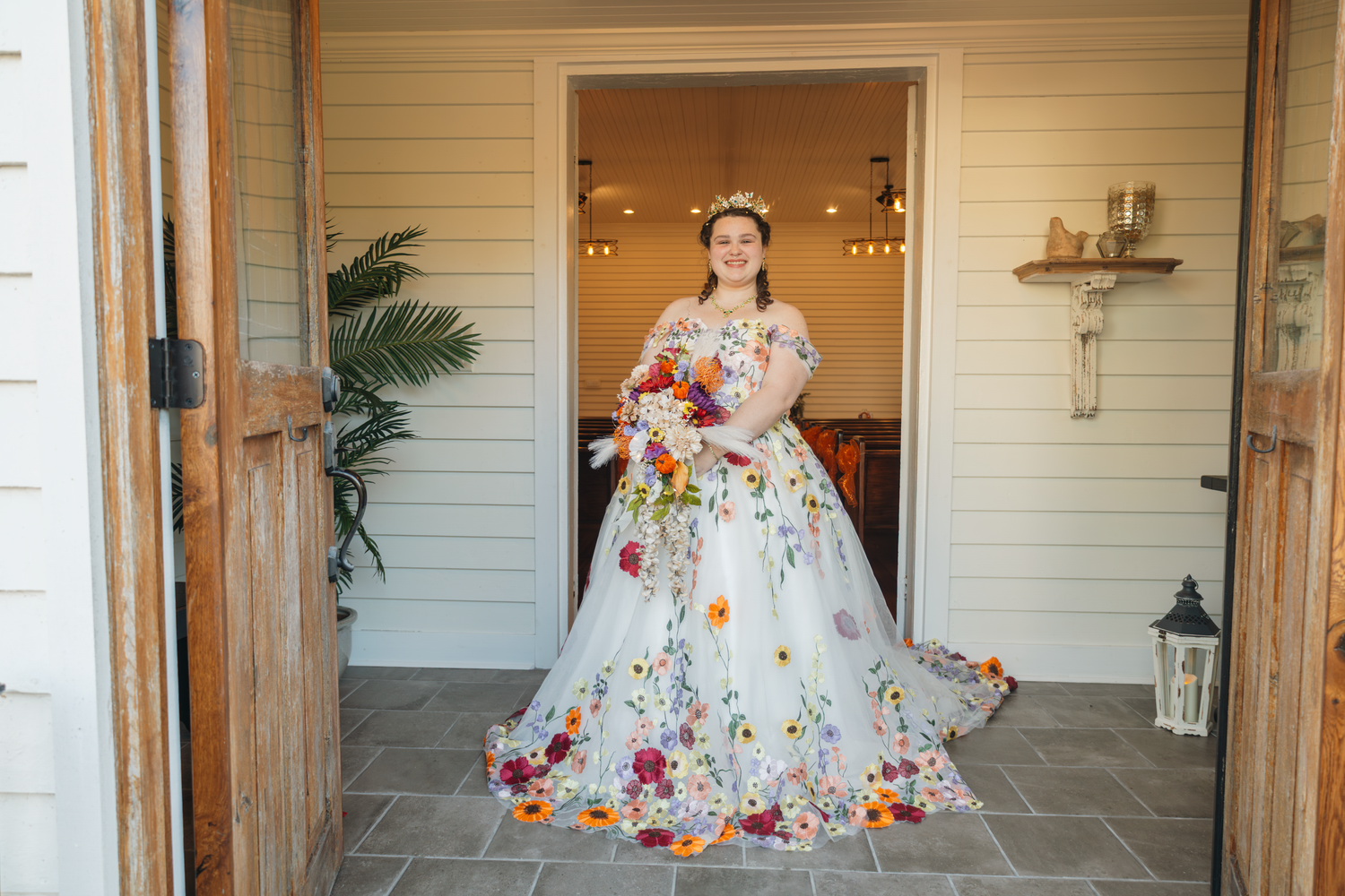 Bride in floral wedding gown holding colorful bouquet stands in doorway of rustic venue.