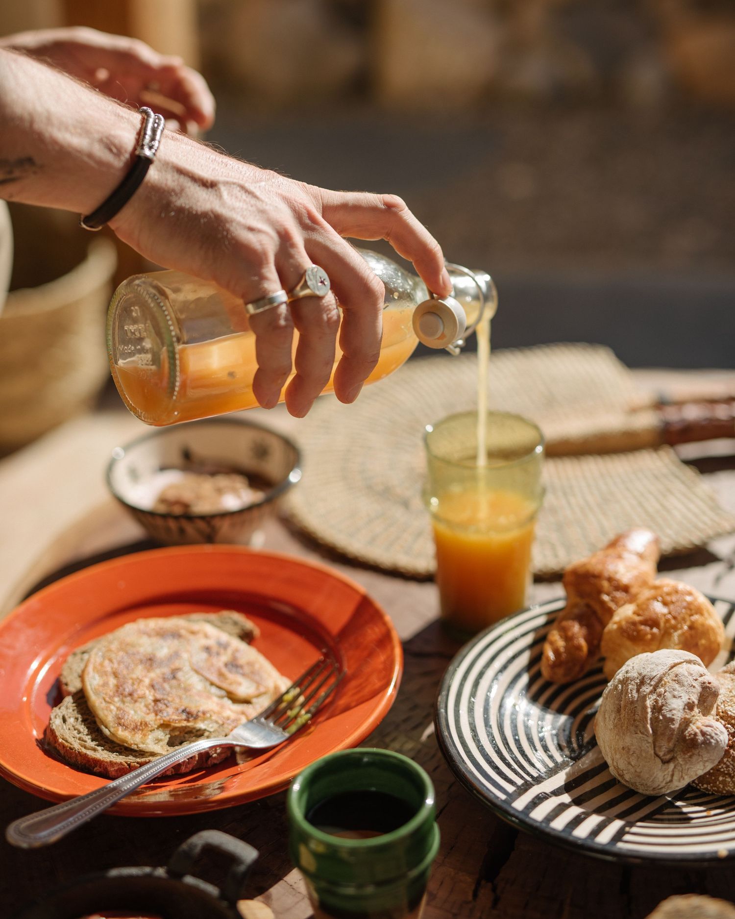 Hands pouring orange juice and serving breakfast items on striped plates during a morning meal setting.