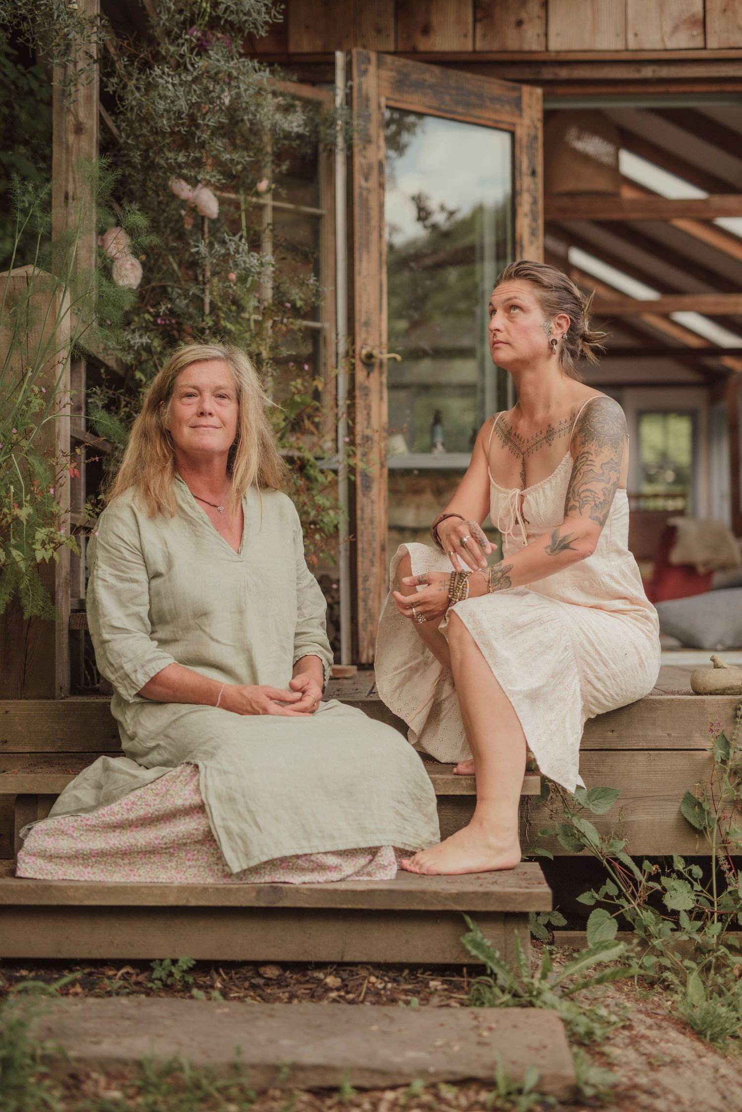 Two people in linen dresses enjoy tea while sitting on rustic wooden porch steps of a cabin.
