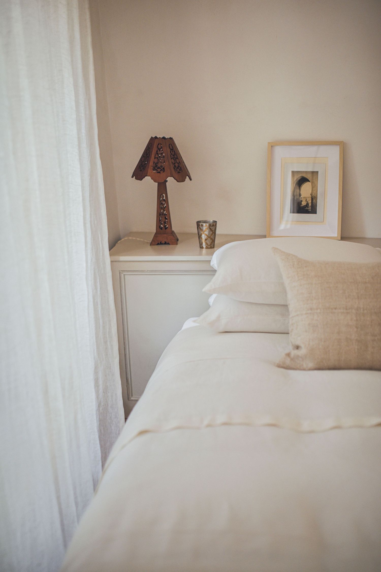 Cozy bedroom corner with white bedding, decorative lamp and framed artwork on bedside table.