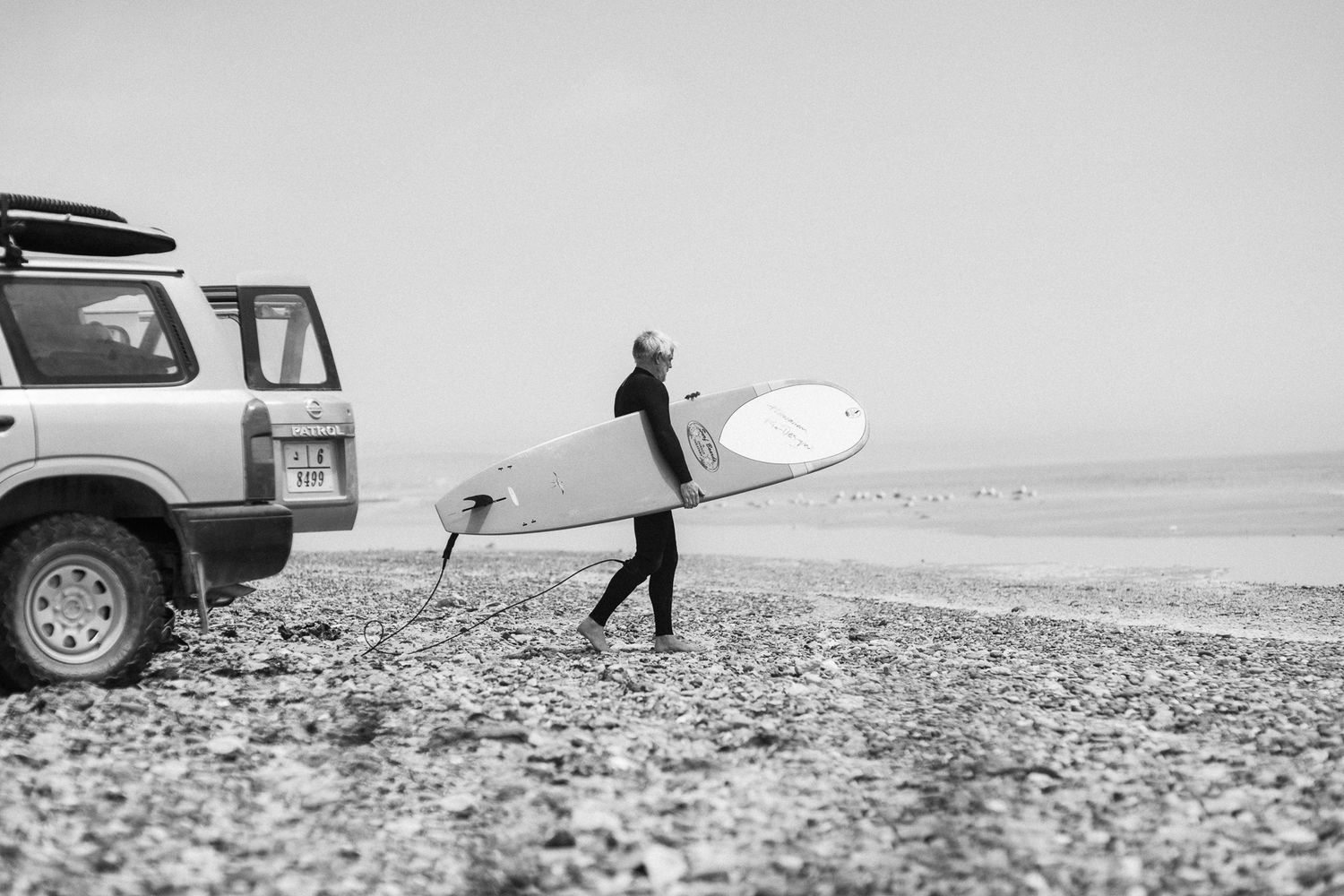 A surfer in a wetsuit carries a white surfboard near a parked 4x4 vehicle on a rocky beach in black and white.