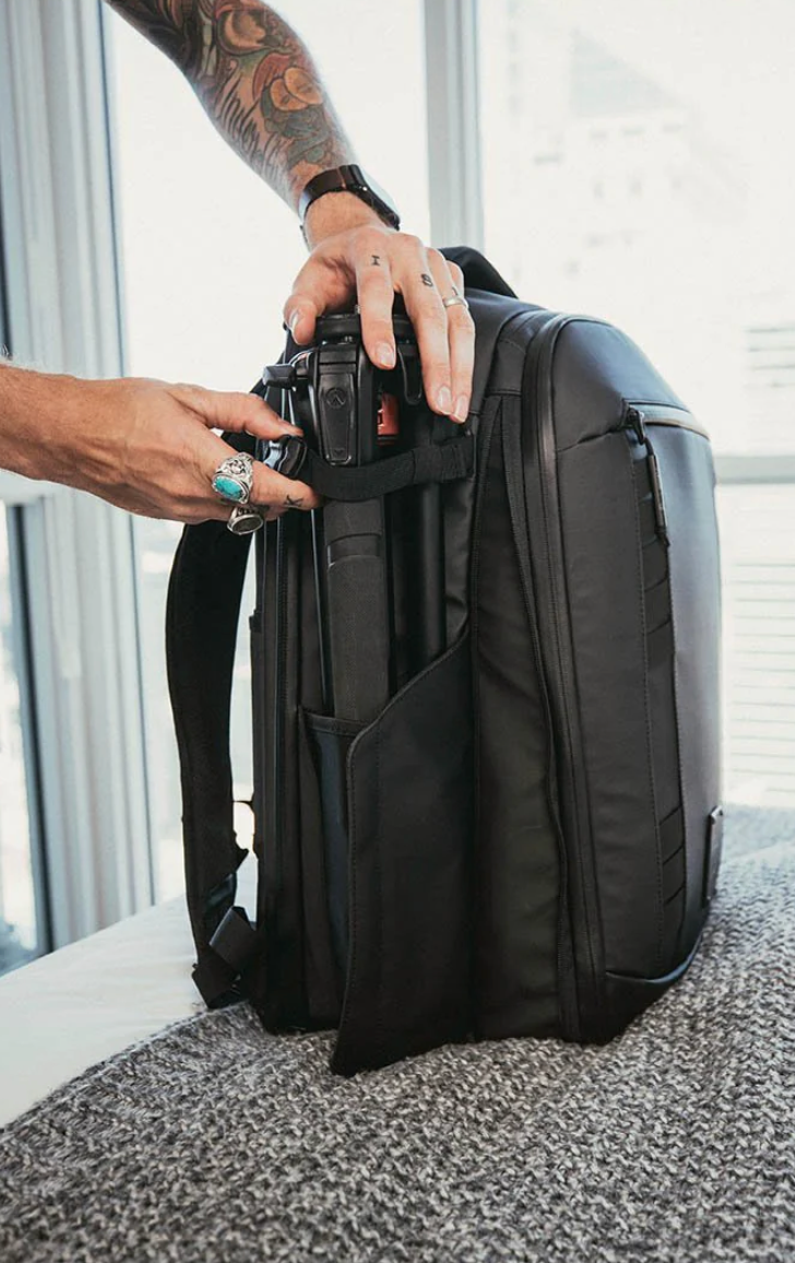 Tattooed hands opening a large black camera backpack on carpet.