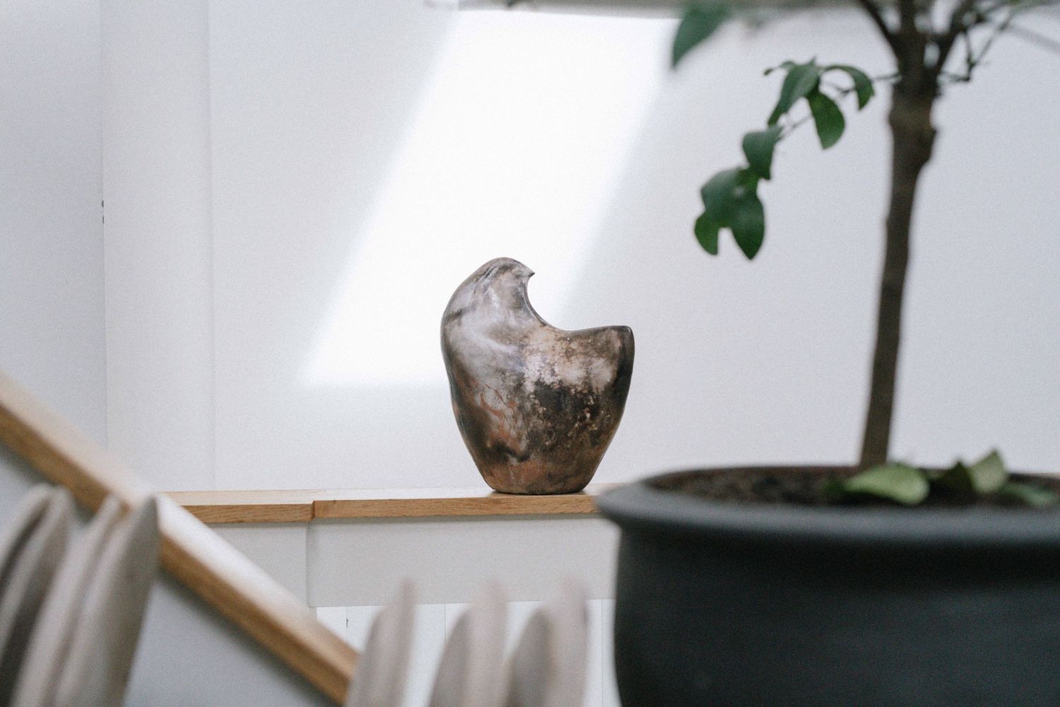 A rustic ceramic vase sits on a wooden ledge near a potted plant in natural indoor lighting.