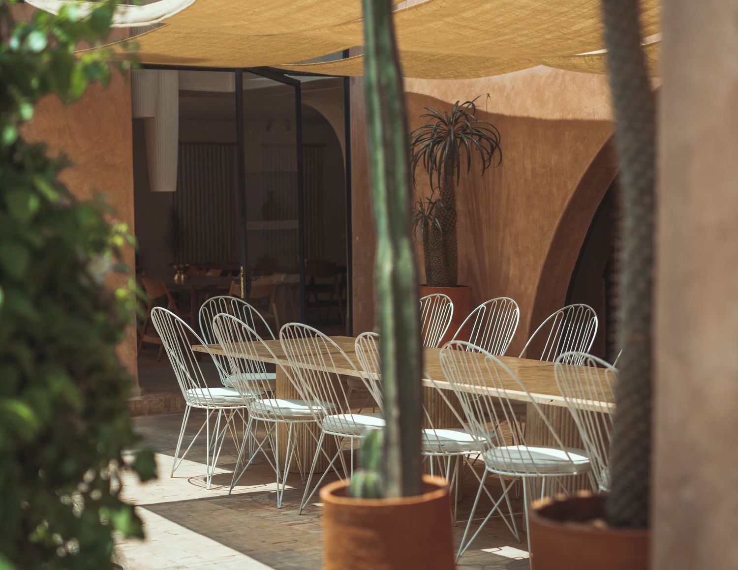 Outdoor patio dining area with white metal chairs arranged under beige awnings and lush green plants.