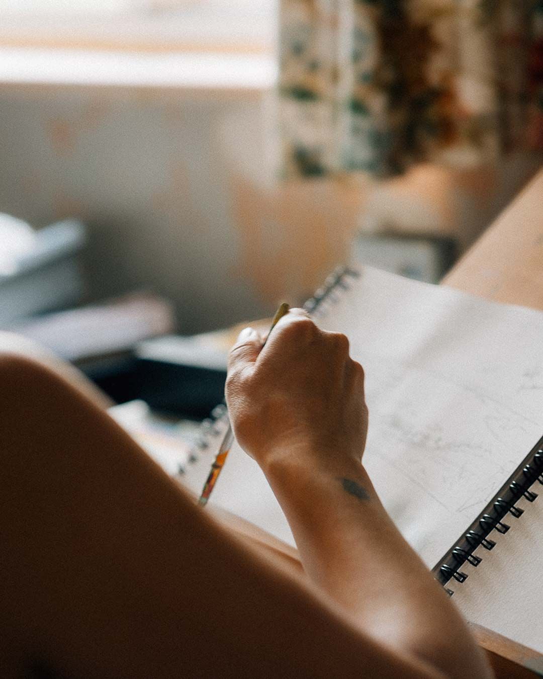 A person writing in a spiral notebook with a pencil, captured in warm natural light near a window.