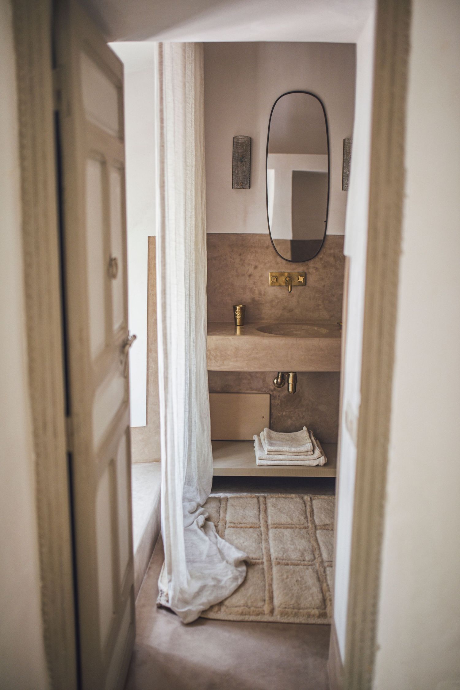 Bathroom doorway view showing oval mirror and stone sink in neutral tones.