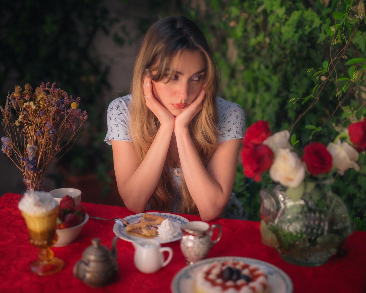 Young woman resting chin on hands at a red table with roses, pastries, and strawberries in a garden setting.