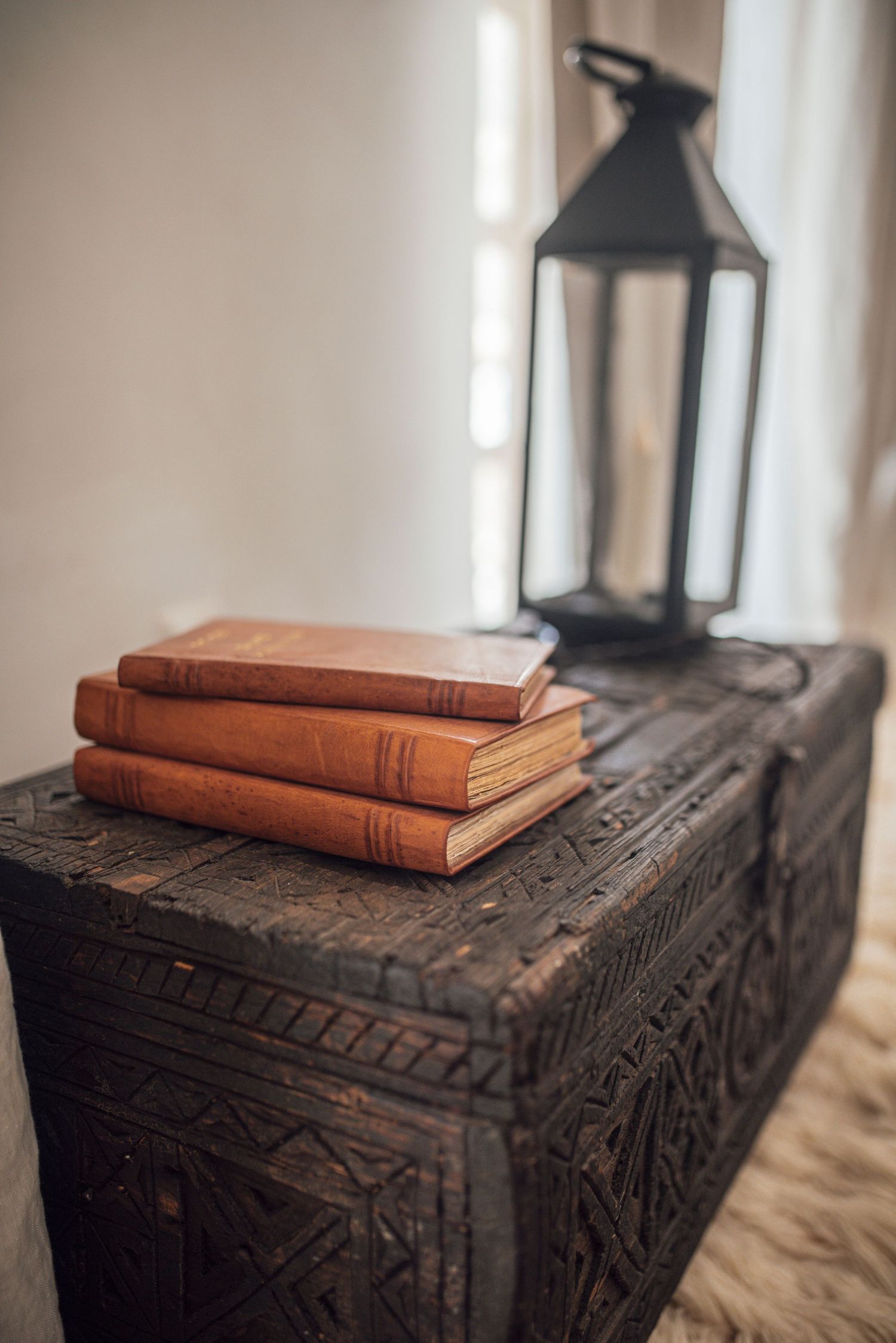 Vintage leather books stacked on carved wooden chest with black lantern in soft window light.