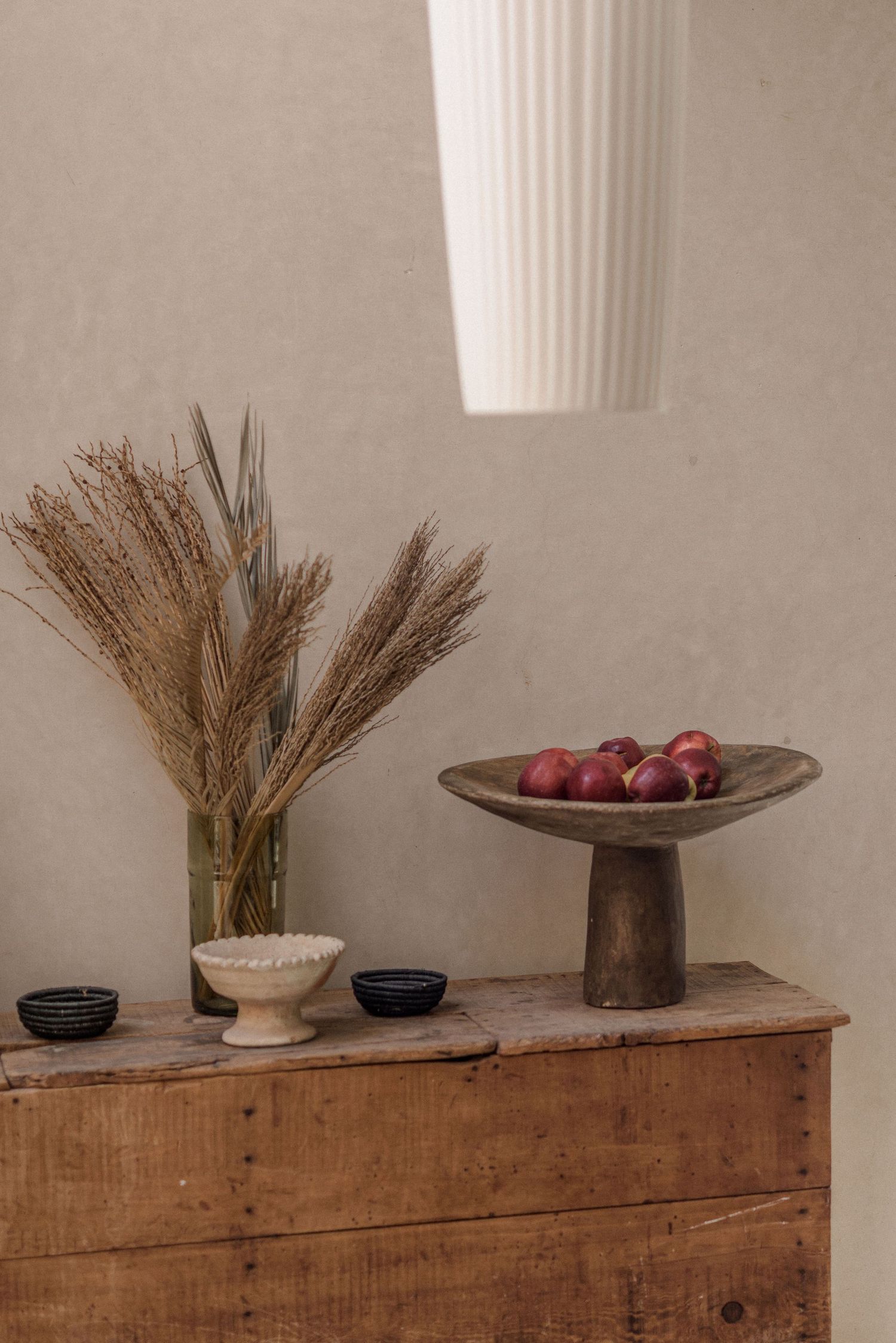 Minimalist still life series showing dried wheat on rustic wooden cabinet with pottery and fruit in warm tones.