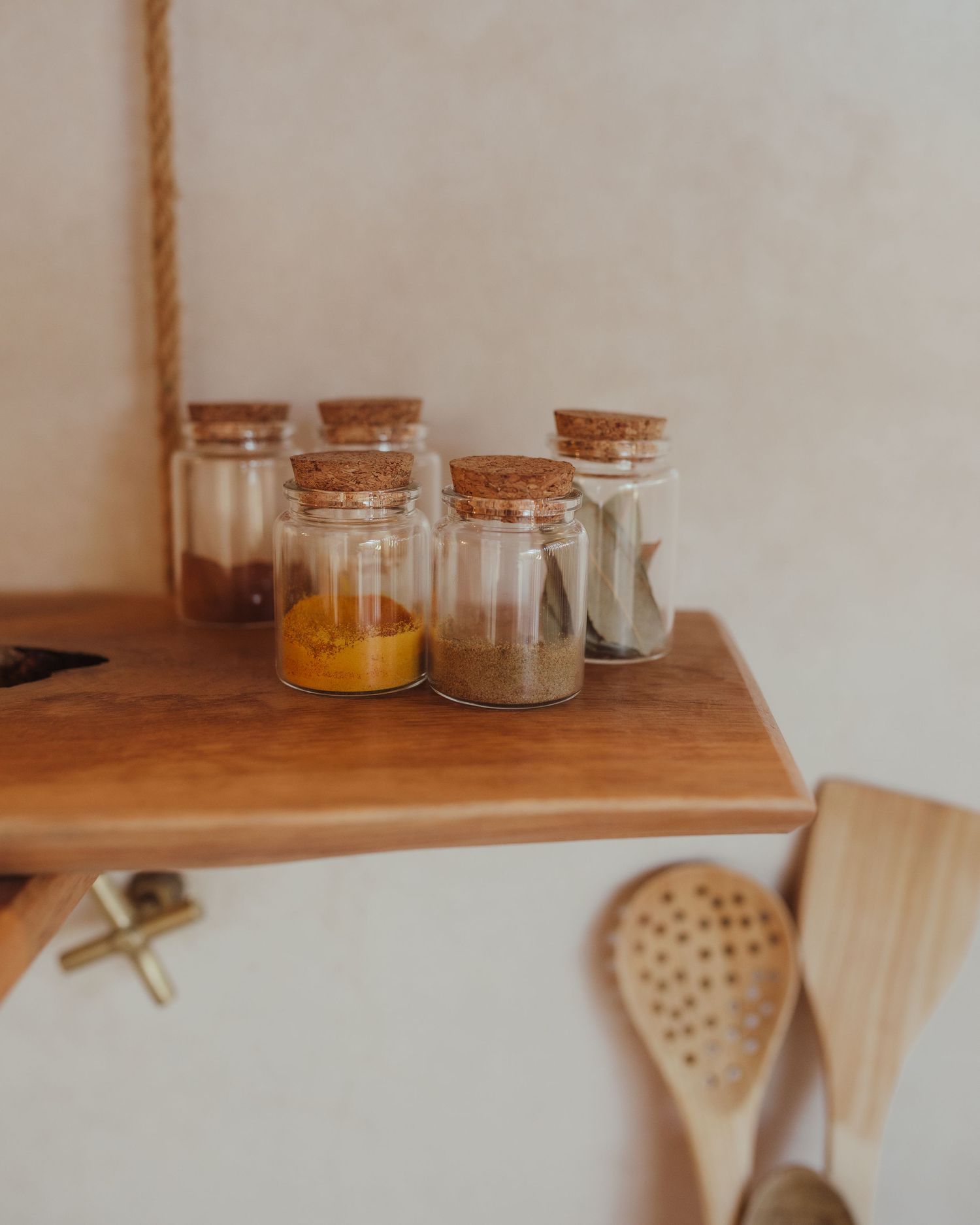 Small glass spice jars with cork lids arranged on a wooden shelf next to kitchen utensils.