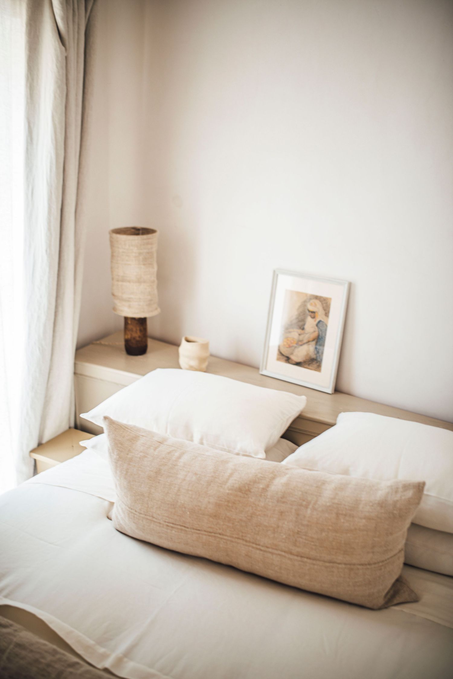 Minimalist bedroom with white linens, beige pillows and neutral decor on bedside table.