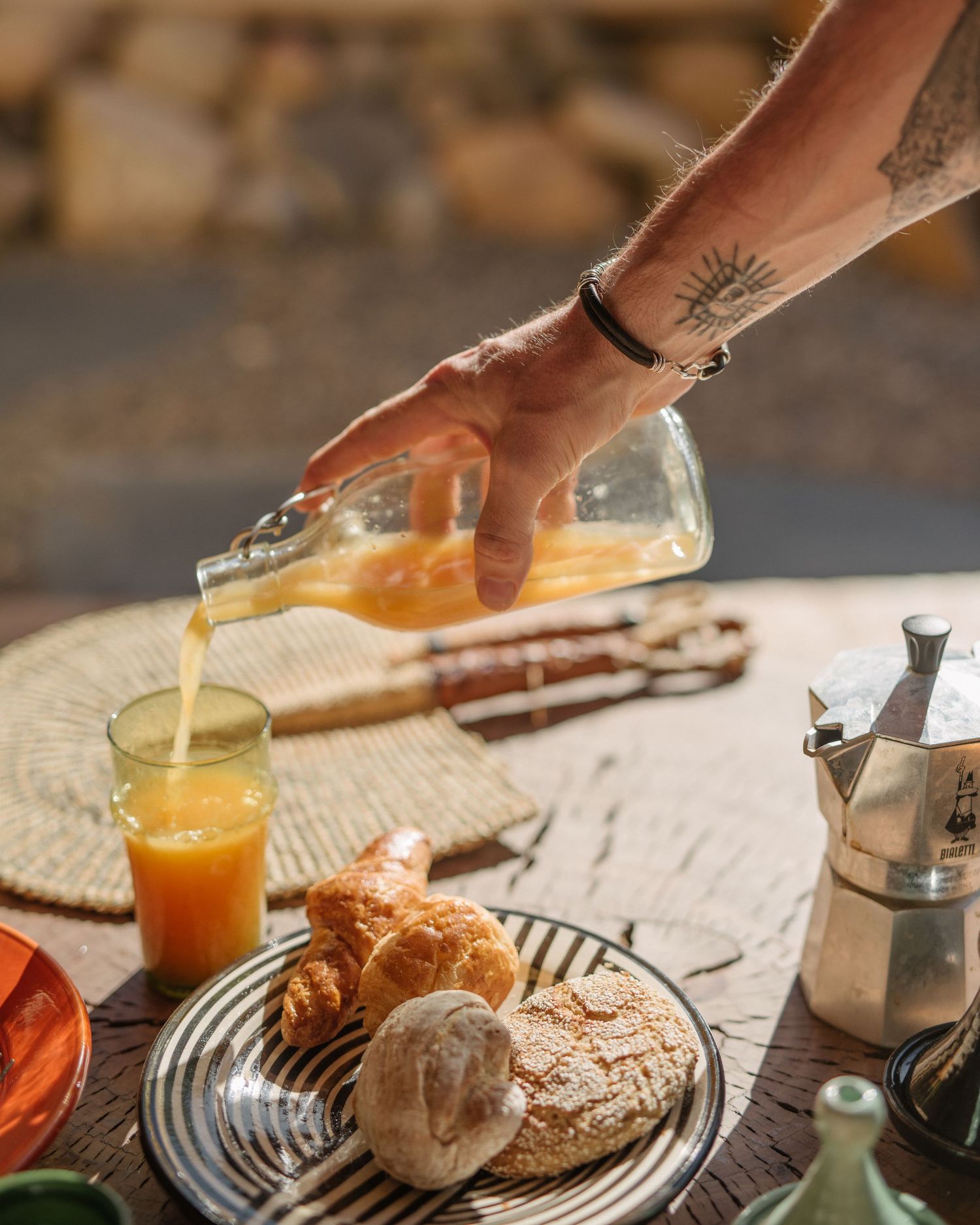 Orange juice being poured into a glass next to fresh croissants and bread on a round serving plate.