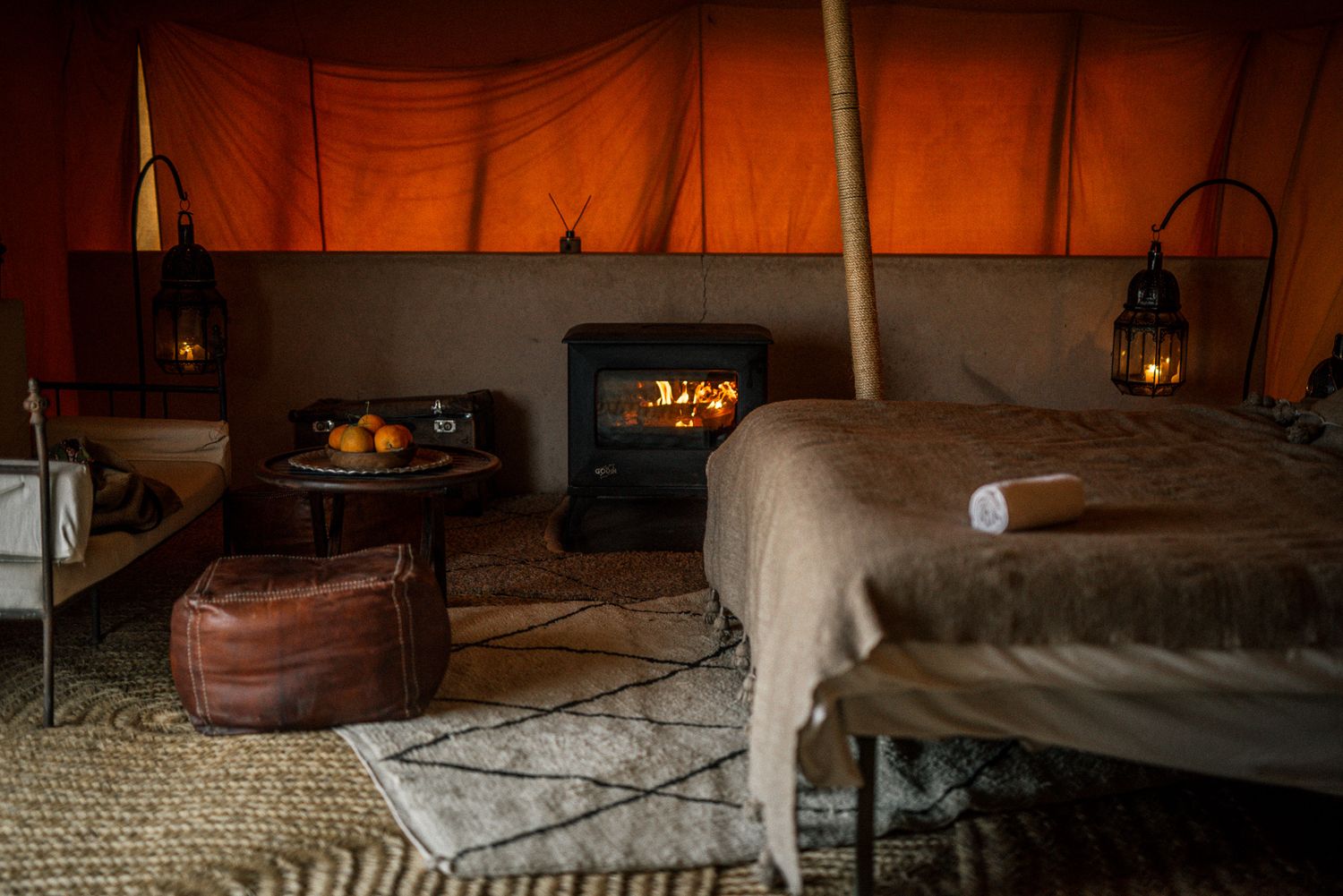 Cozy glamping tent interior with wood stove, lanterns and plush bedding in warm ambient lighting.