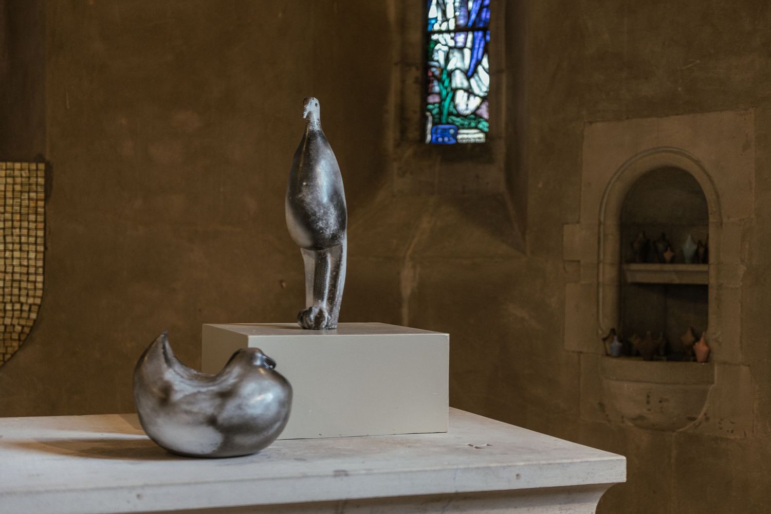Artistic sculptures displayed on white pedestals in a dimly lit gallery space with stone walls and arched windows.