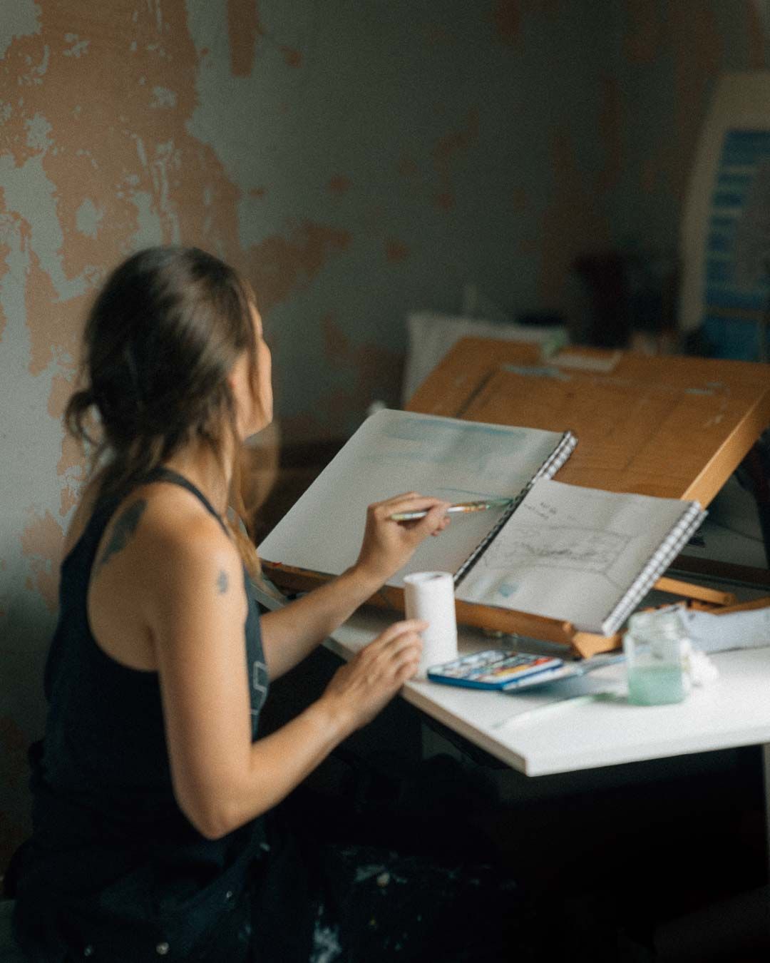 Person wearing dark clothing sits at desk studying textbooks and taking notes in dim lighting.