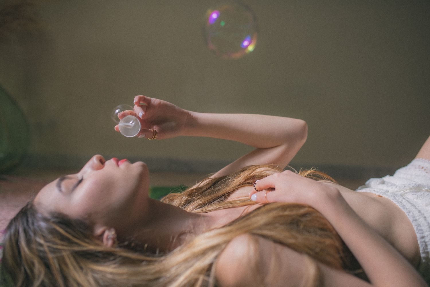 Young woman lying down blowing soap bubbles, holding bubble wand with long blonde hair spread out.