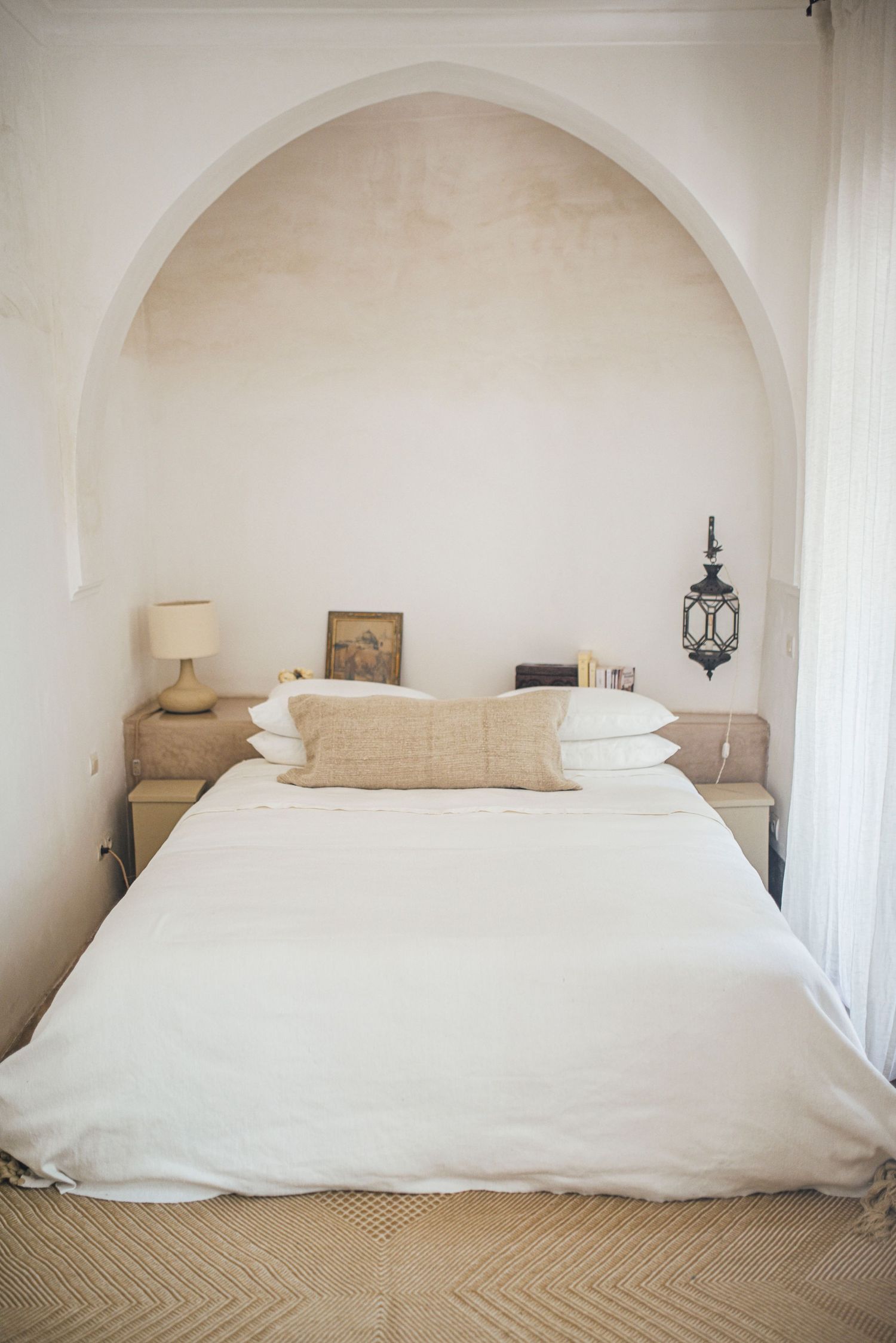 Minimalist bedroom with arched alcove, white bedding, and Moroccan-style lantern.