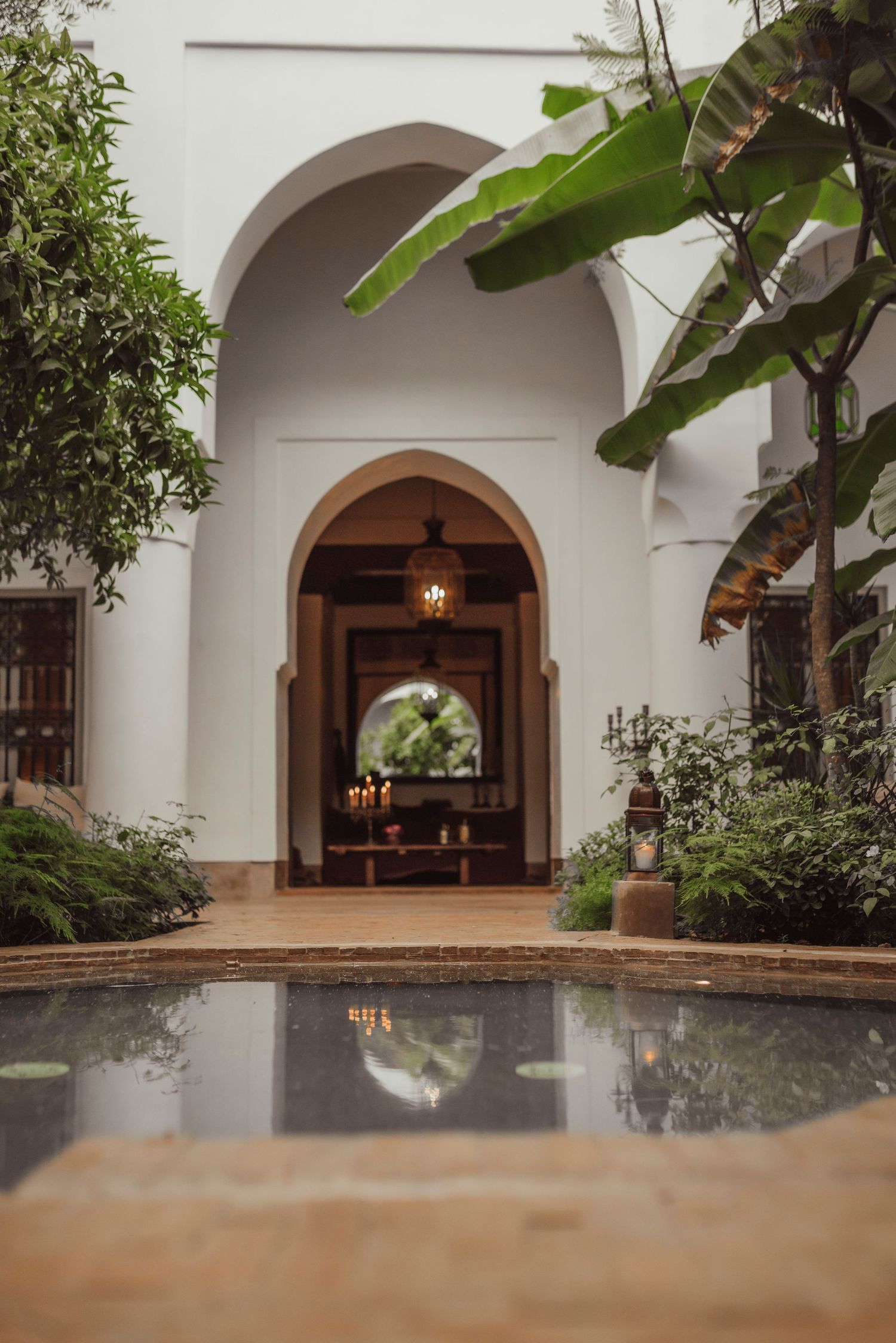 Elegant arched walkway with tropical plants and reflecting pool leading to illuminated doorway.
