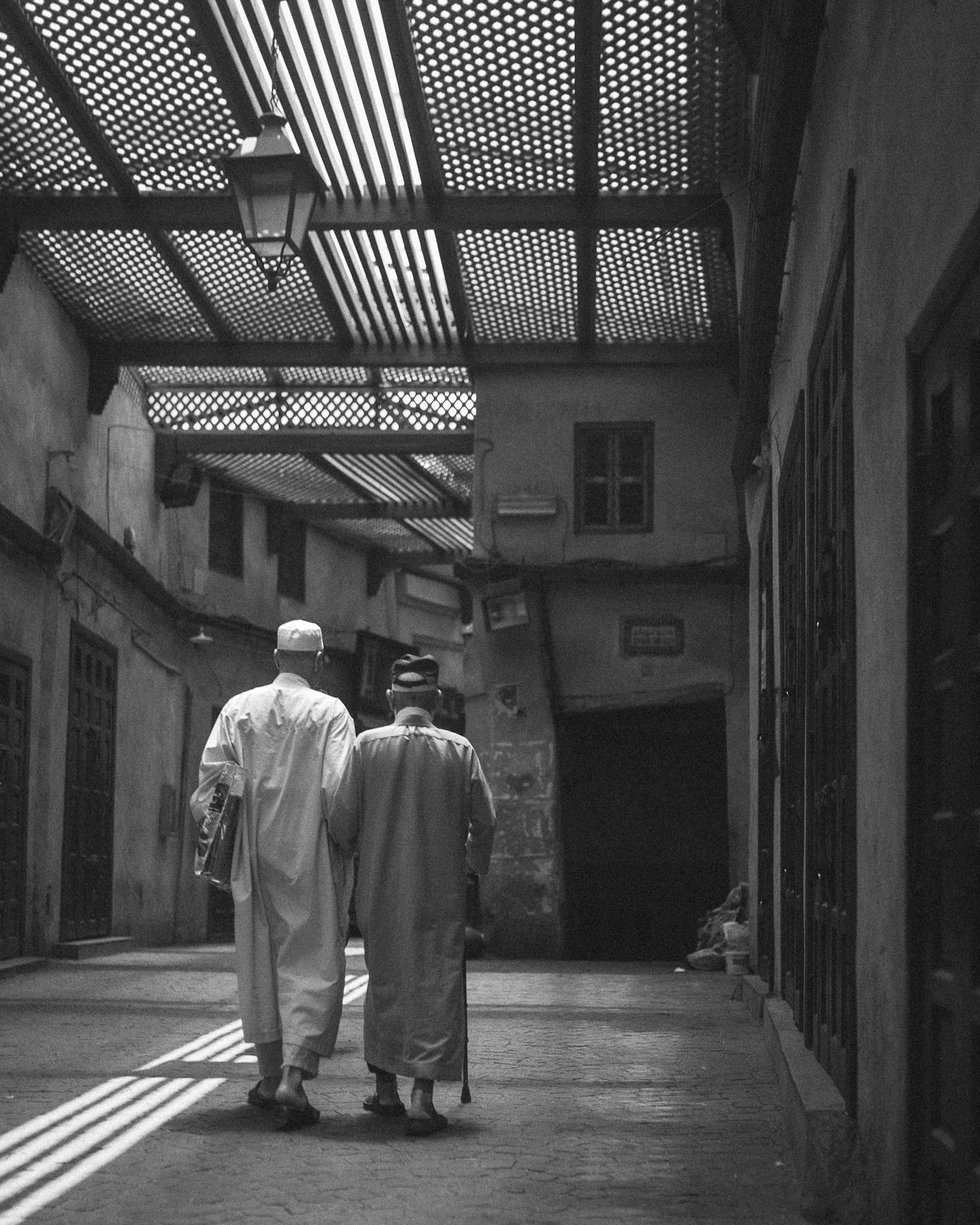 Two figures in traditional robes walk together under a latticed roof casting dramatic shadows in a narrow alleyway.