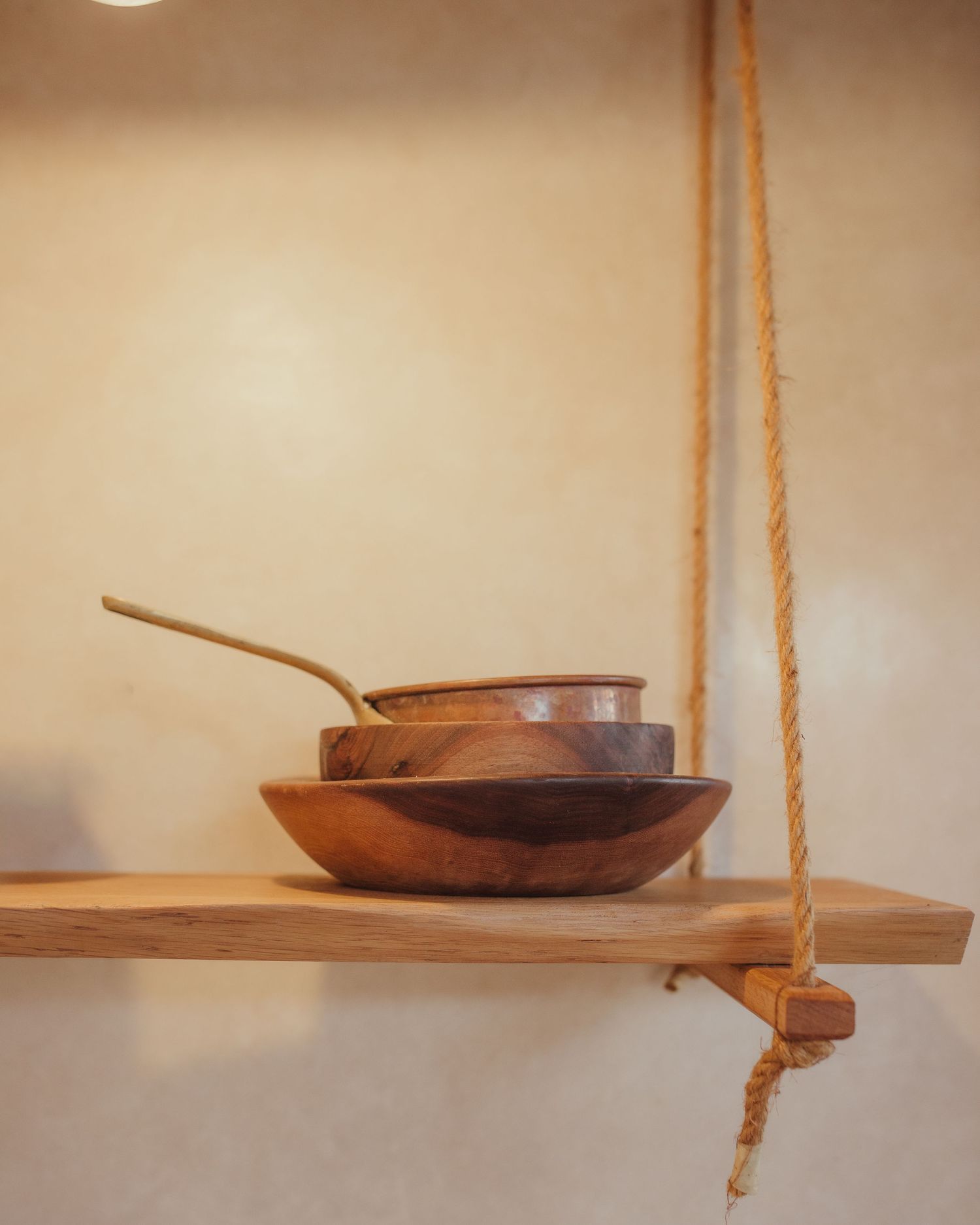 Wooden bowls and pans arranged on a rope-suspended floating shelf against a beige wall.