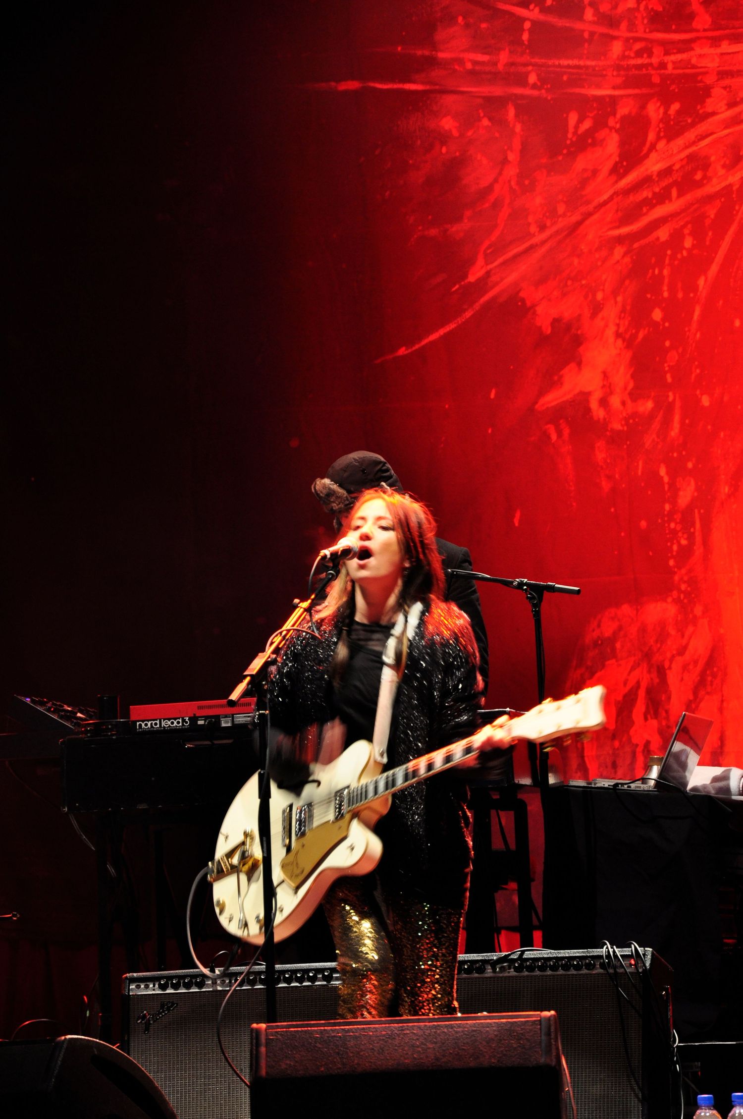 A performer plays guitar and sings on stage bathed in dramatic red lighting effects during a live concert performance.