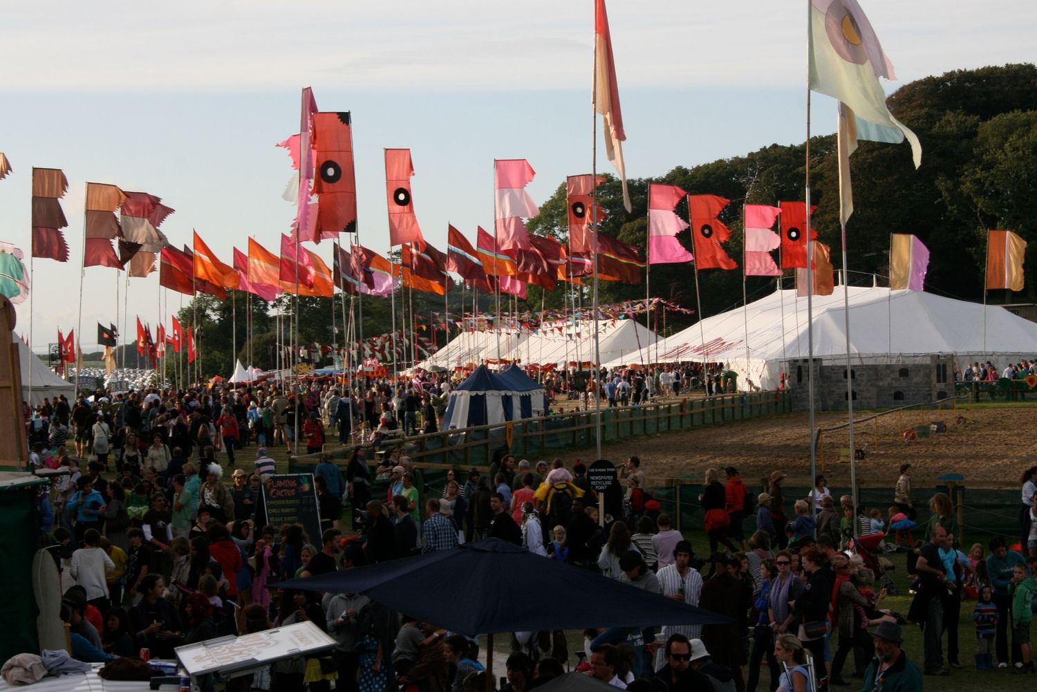 Outdoor festival scene with rows of red flags and white tents stretching across a grassy field in daylight.