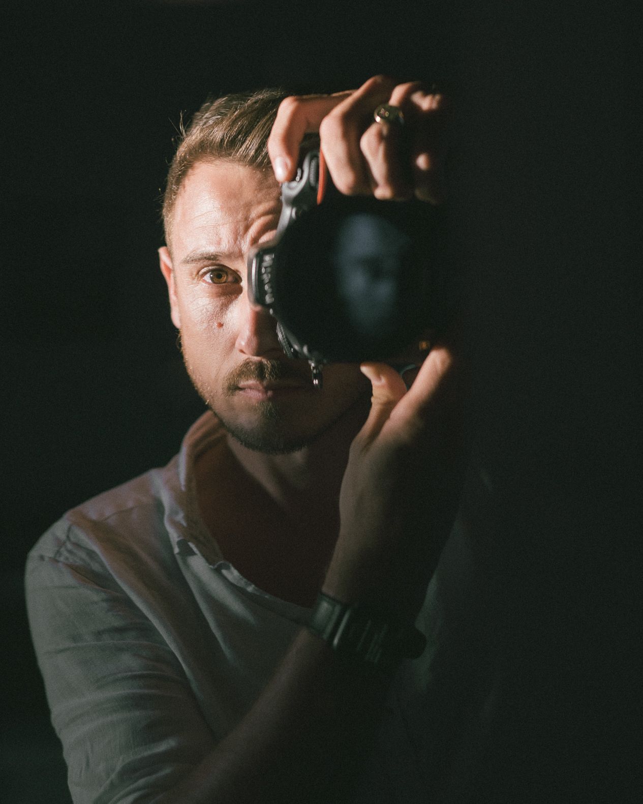 A photographer holds up a camera in a dark studio, with dramatic side lighting creating a moody self-portrait.