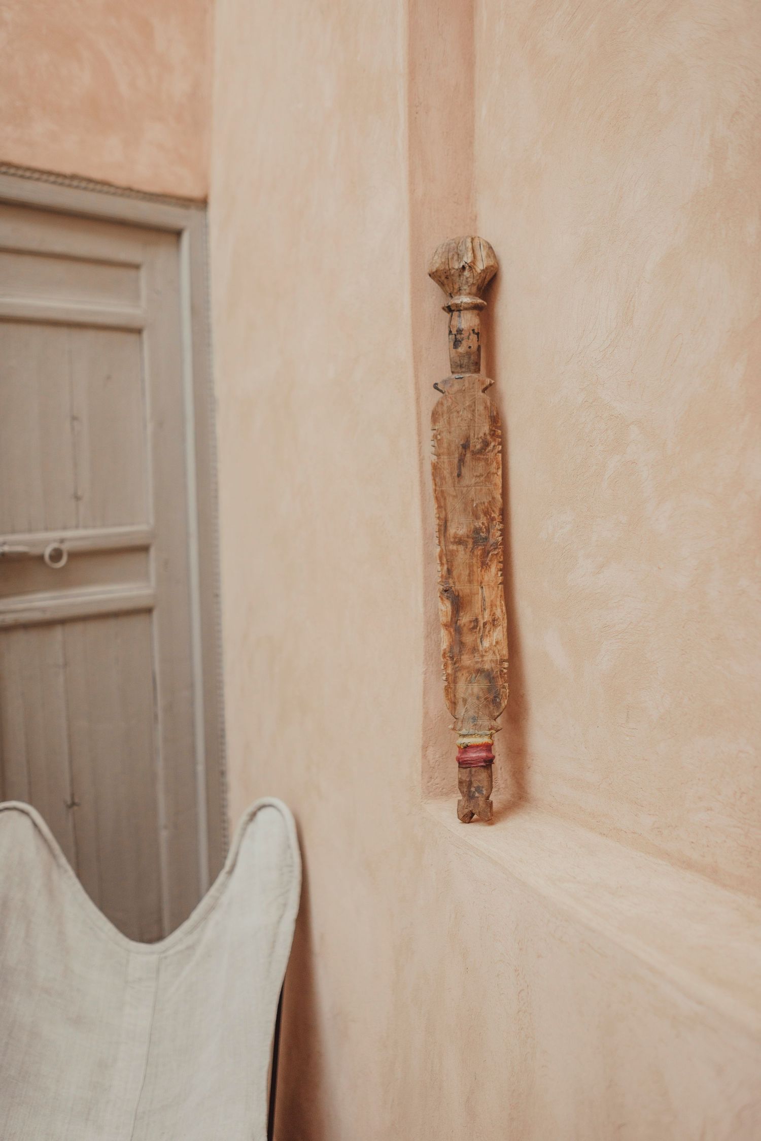 A weathered wooden decorative piece mounted on a beige wall next to a white chair.