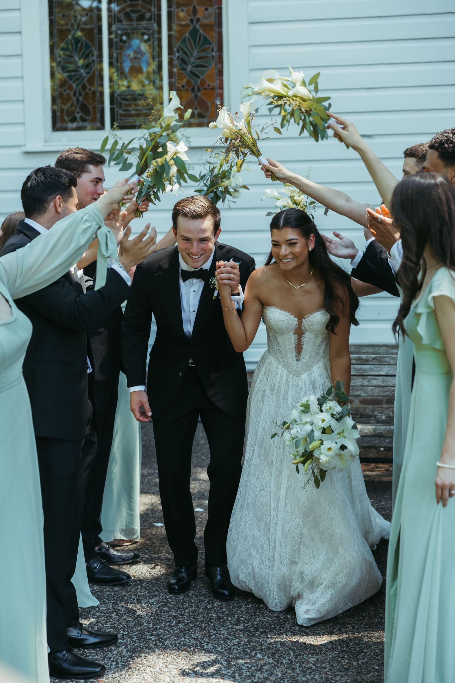 Wedding party celebrates with raised flowers as couple walks through.