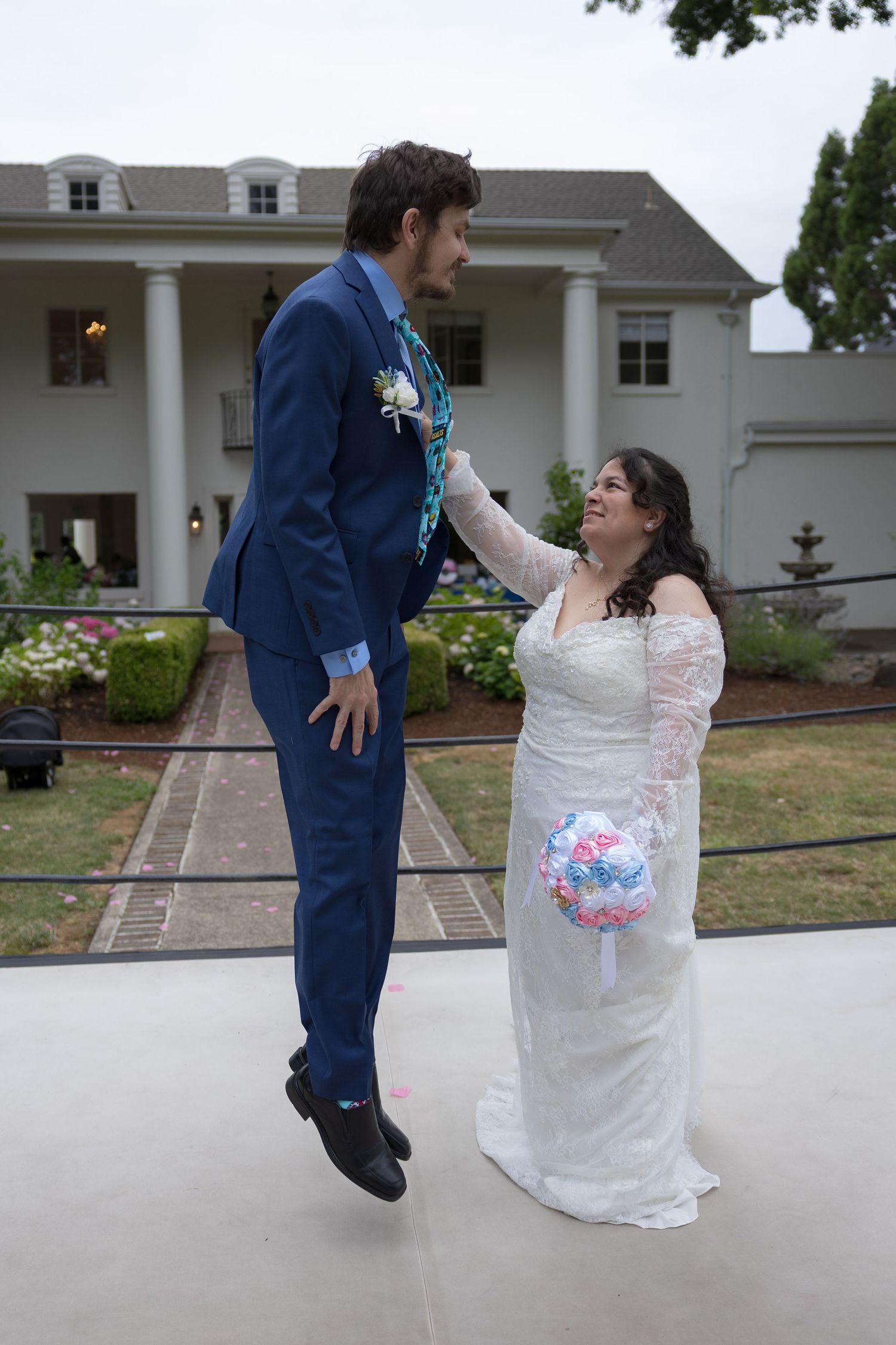 A couple in wedding attire dances together on a paved area in front of a white house during their celebration.