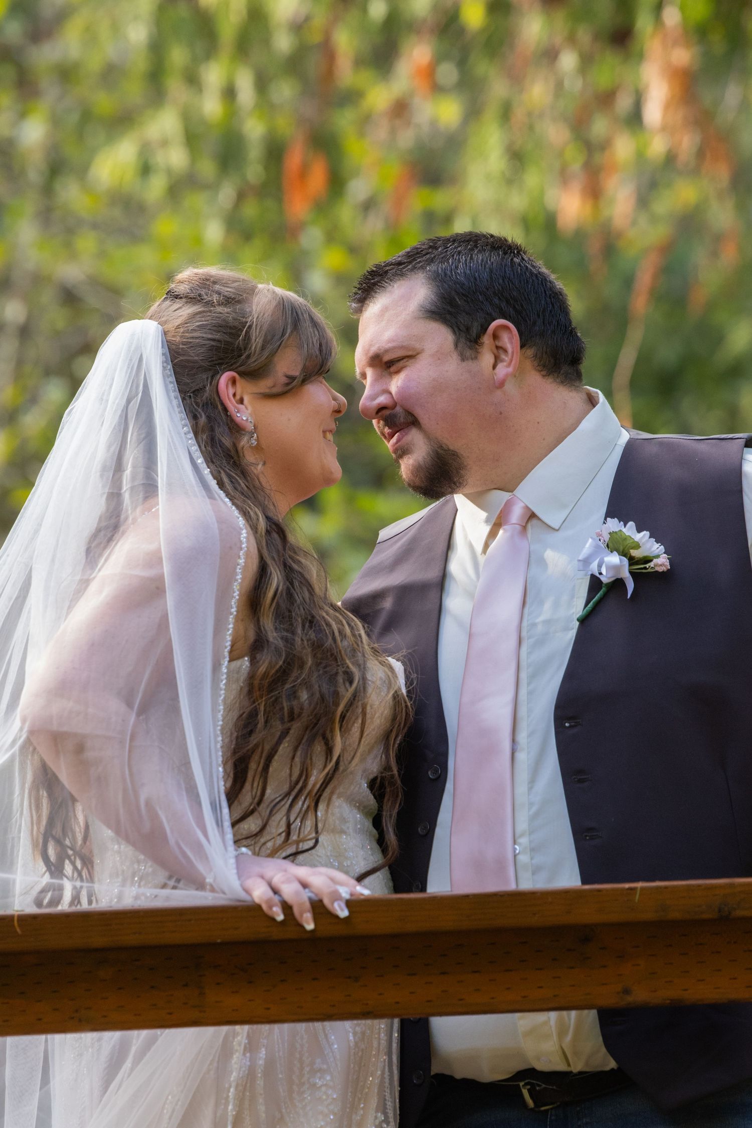 A romantic wedding moment captured outdoors with a bride in white dress and veil leaning close to her groom in a dark suit.