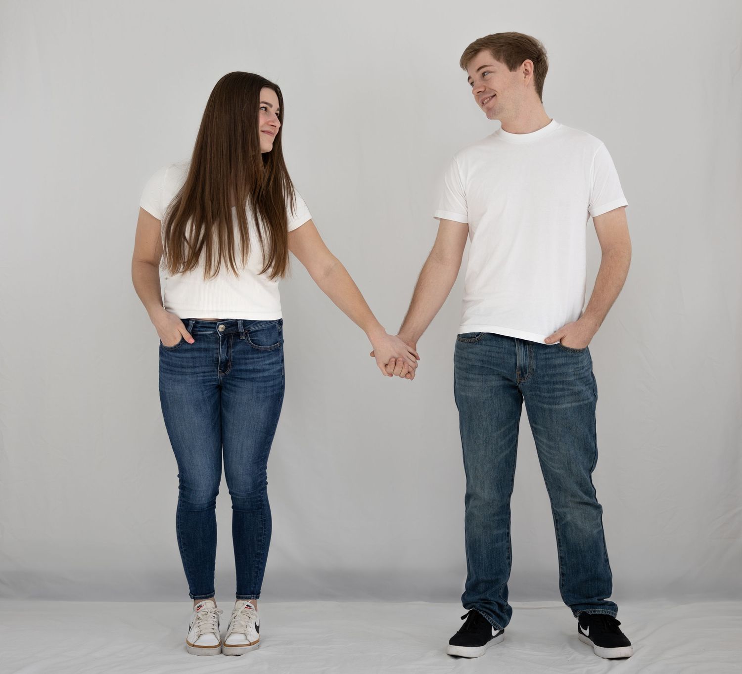 A couple wearing white t-shirts and blue jeans holds hands while standing against a light gray background.