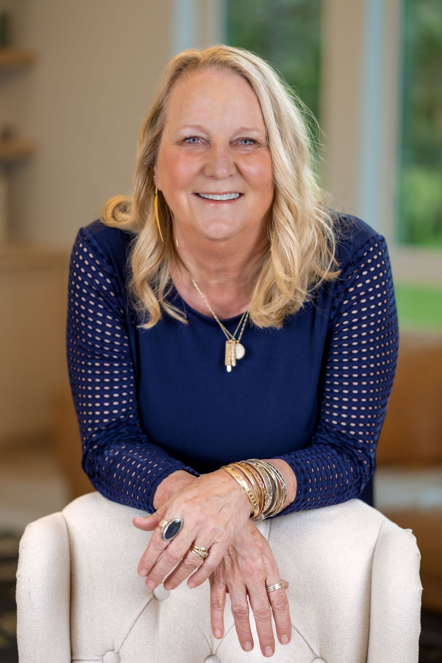 Portrait of someone in a navy blue top with detailed sleeves sitting in a light-filled room.