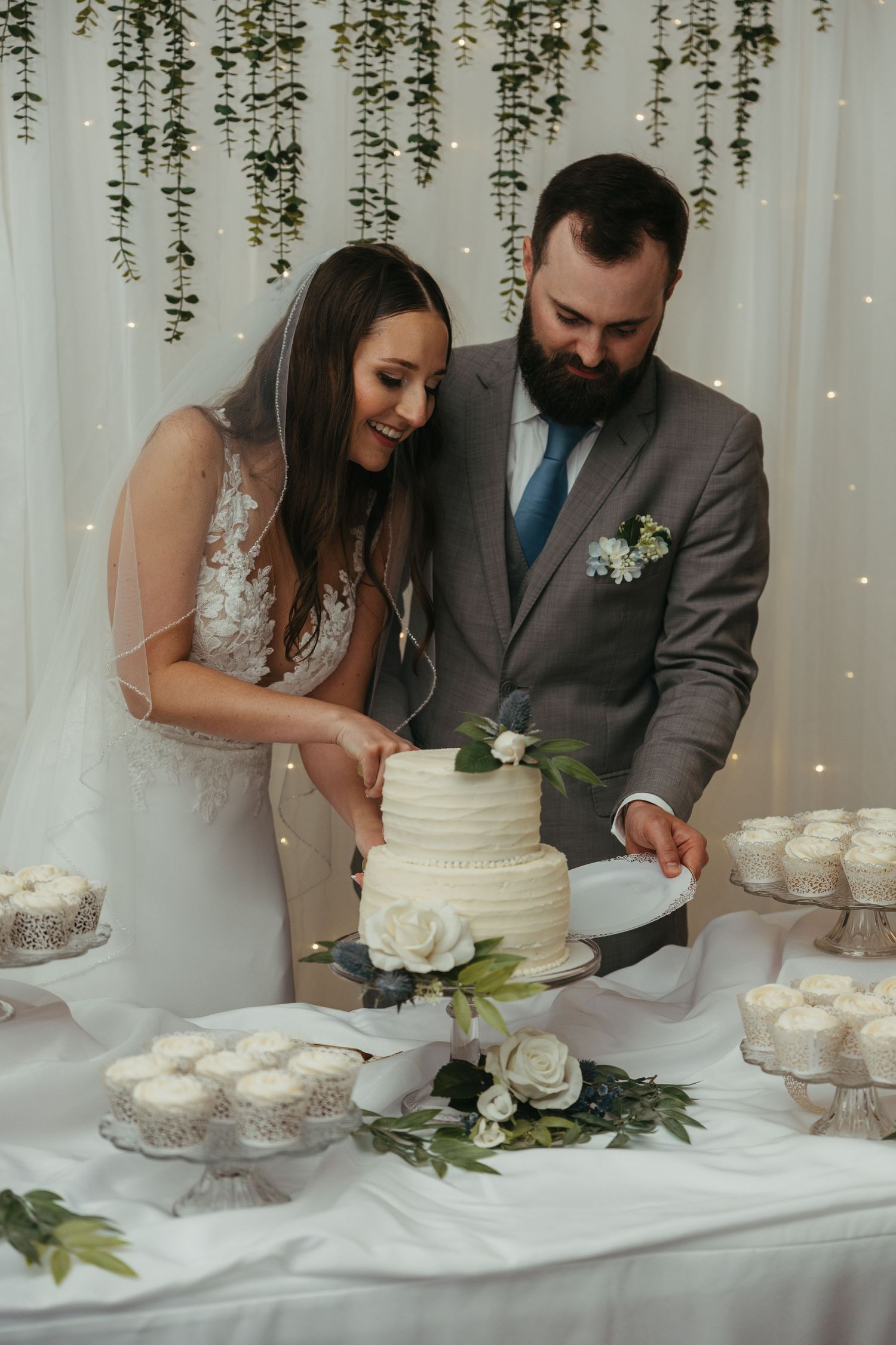 Newlyweds cutting their elegant white wedding cake under hanging greenery decorations.