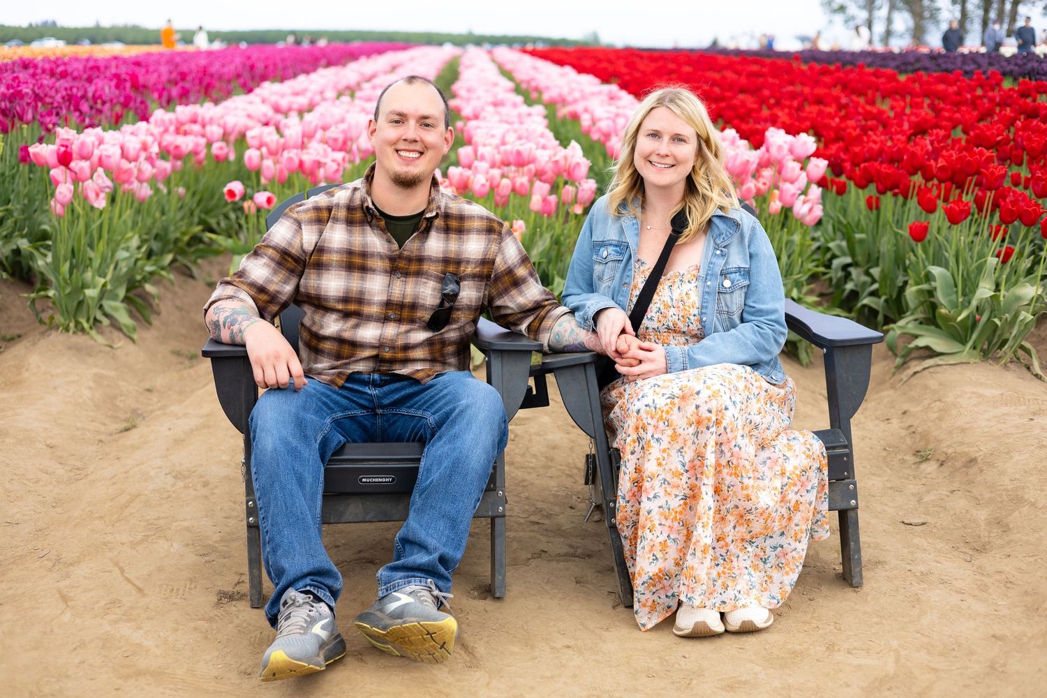 Two people sit in chairs between rows of vibrant pink and red tulips in a flower field.