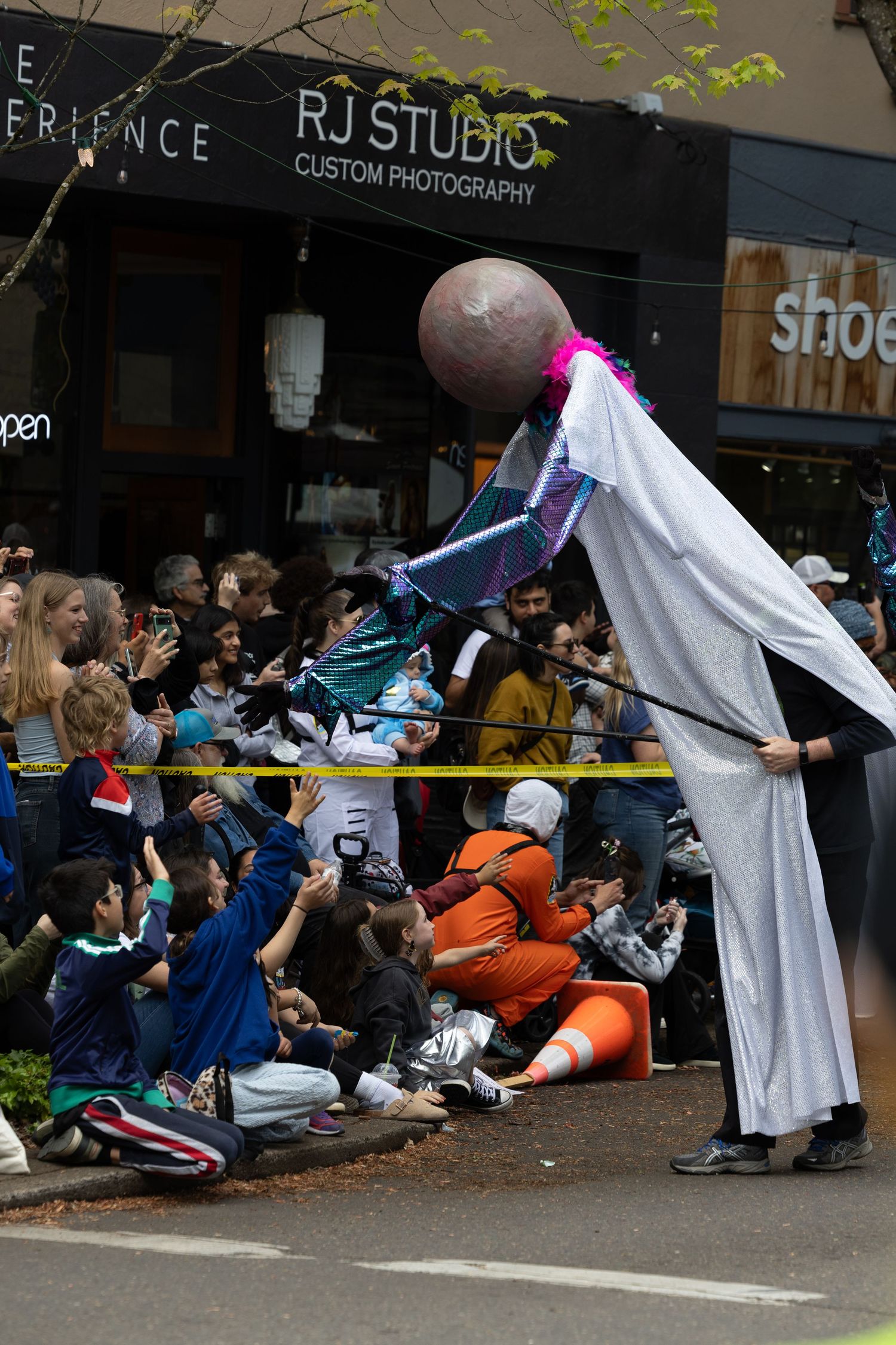 Spectators line the sidewalk watching participants in white robes during an outdoor urban event.