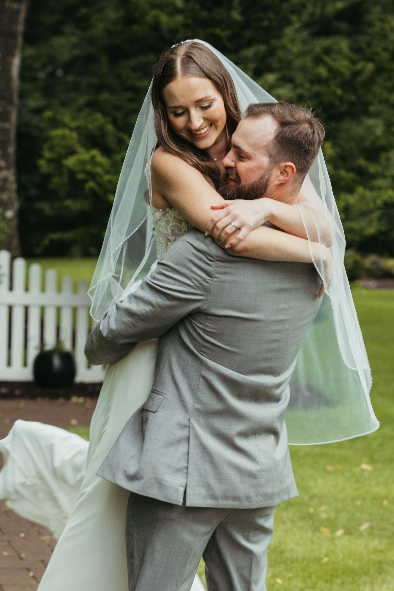 A joyful wedding couple embrace outdoors with the bride in a flowing veil and groom in gray suit.