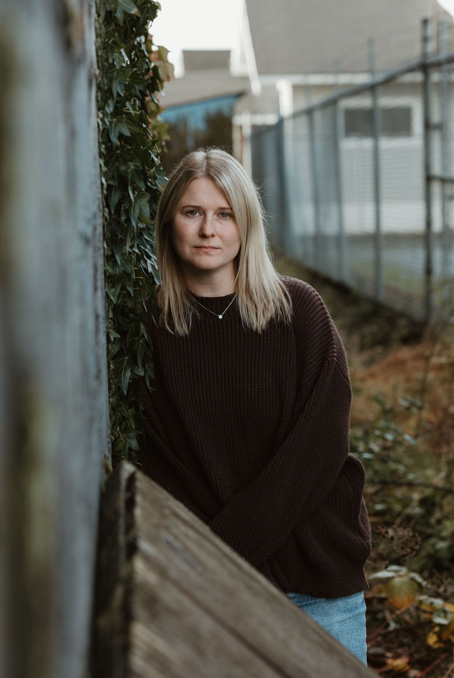 Portrait in earthy tones showing someone wearing a brown sweater against urban greenery.