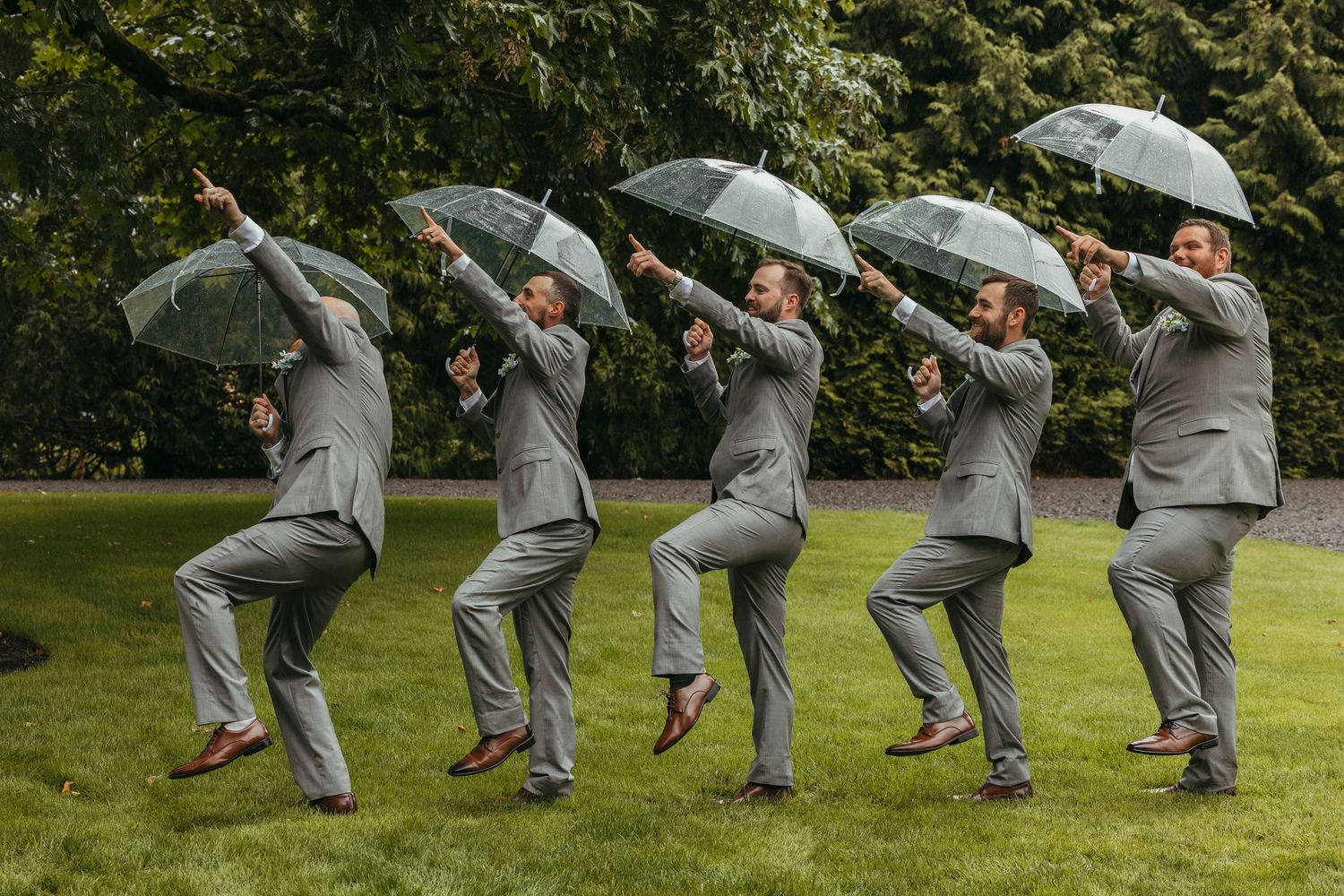 Five groomsmen in grey suits pose playfully with umbrellas on a lawn.
