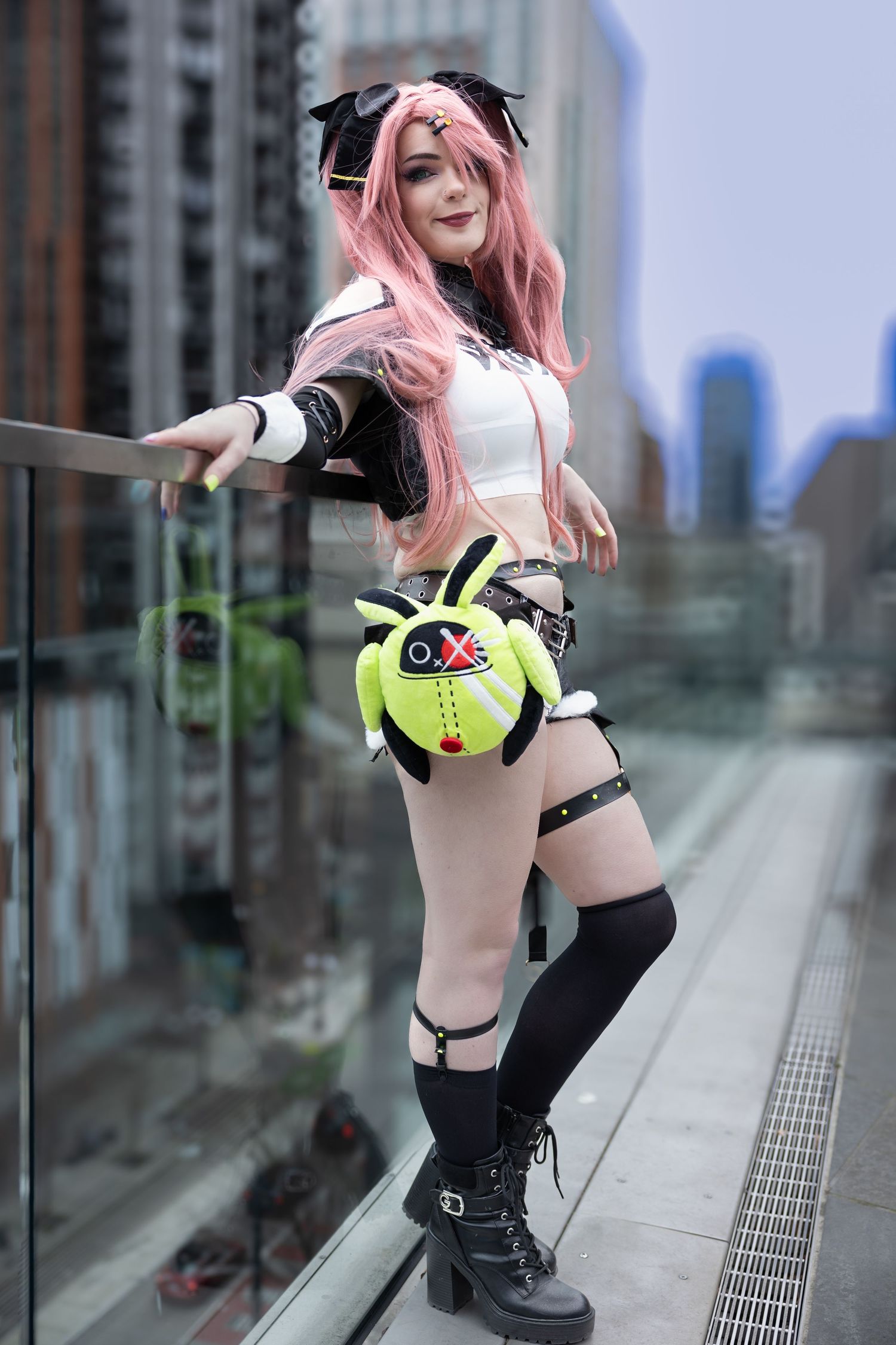 Cosplayer in black and neon yellow cyberpunk outfit poses against urban backdrop with city buildings visible.
