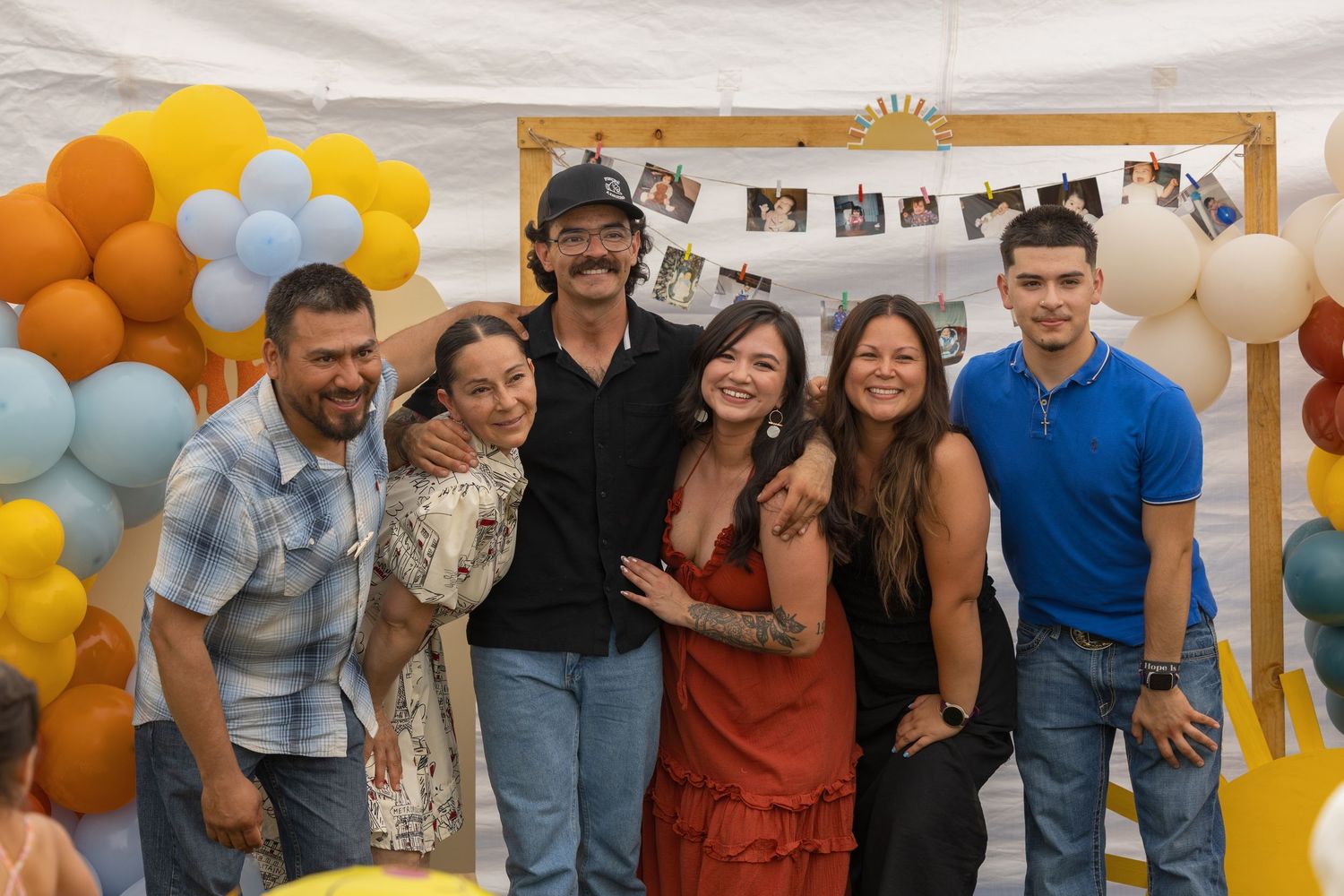 A group celebrates at a party with orange and blue balloon decorations and photos hanging on the wall behind them.