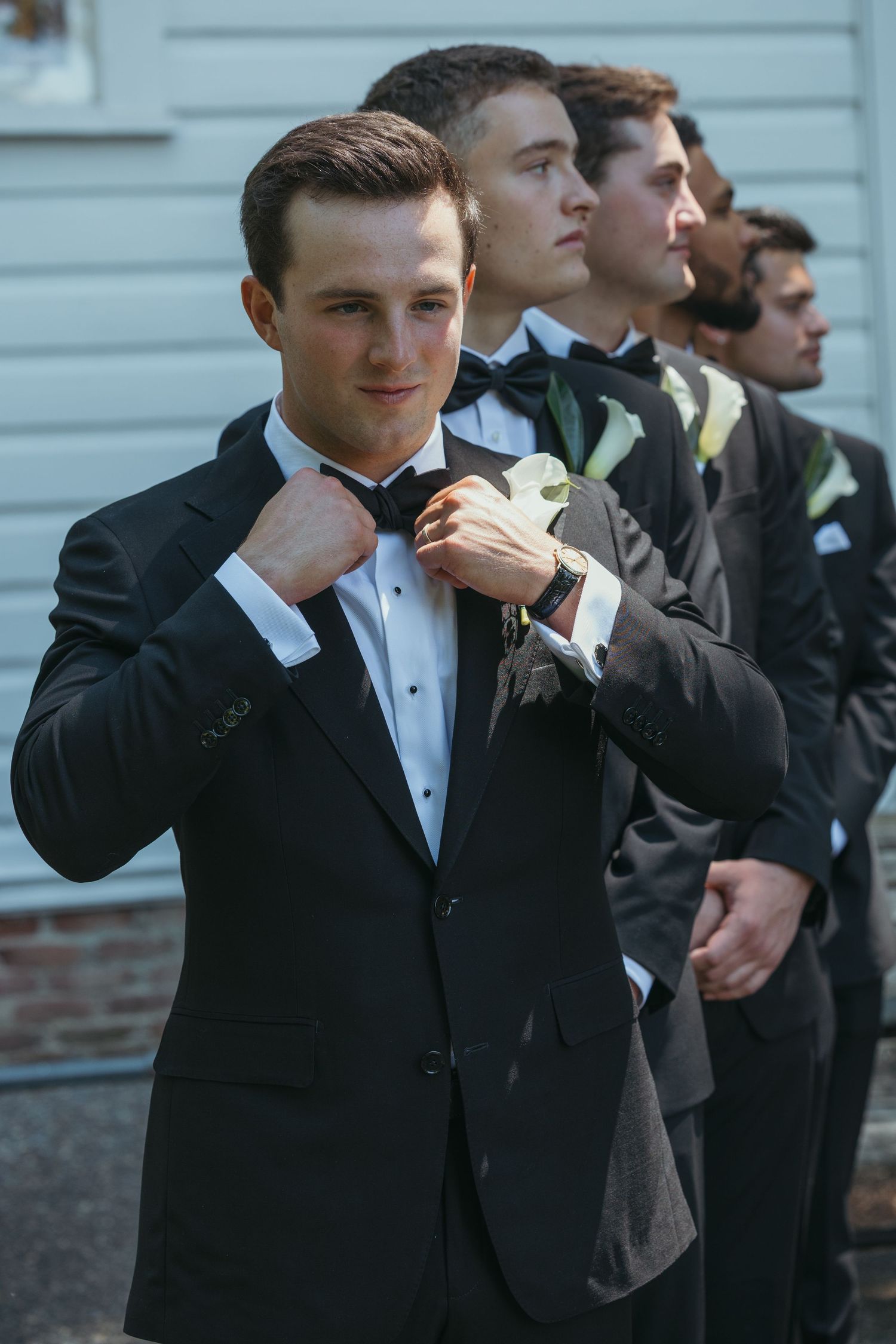 Groomsmen in formal tuxedos adjust their bow ties in synchronized poses.