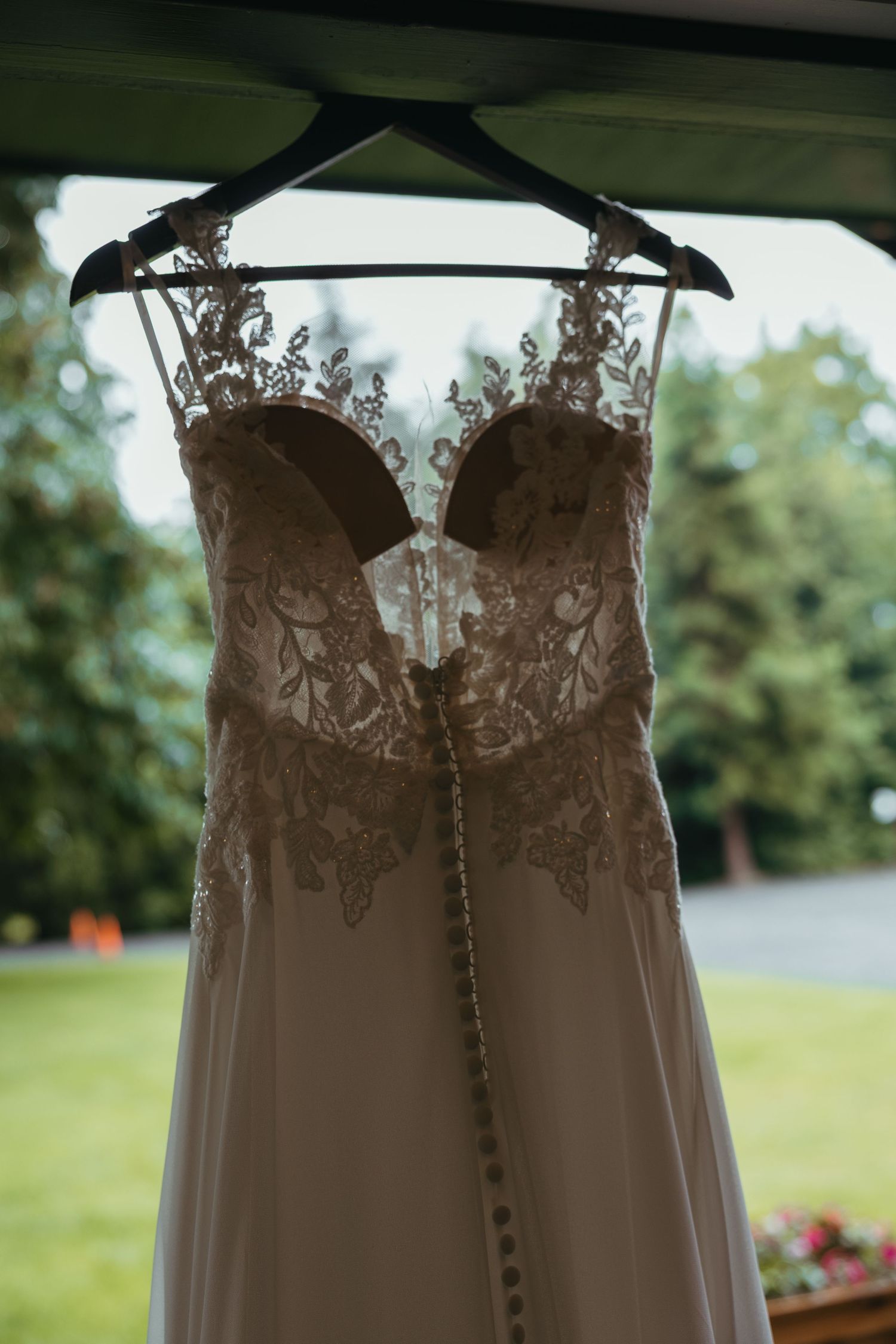 A white wedding dress with intricate lace bodice hangs against green outdoor backdrop.