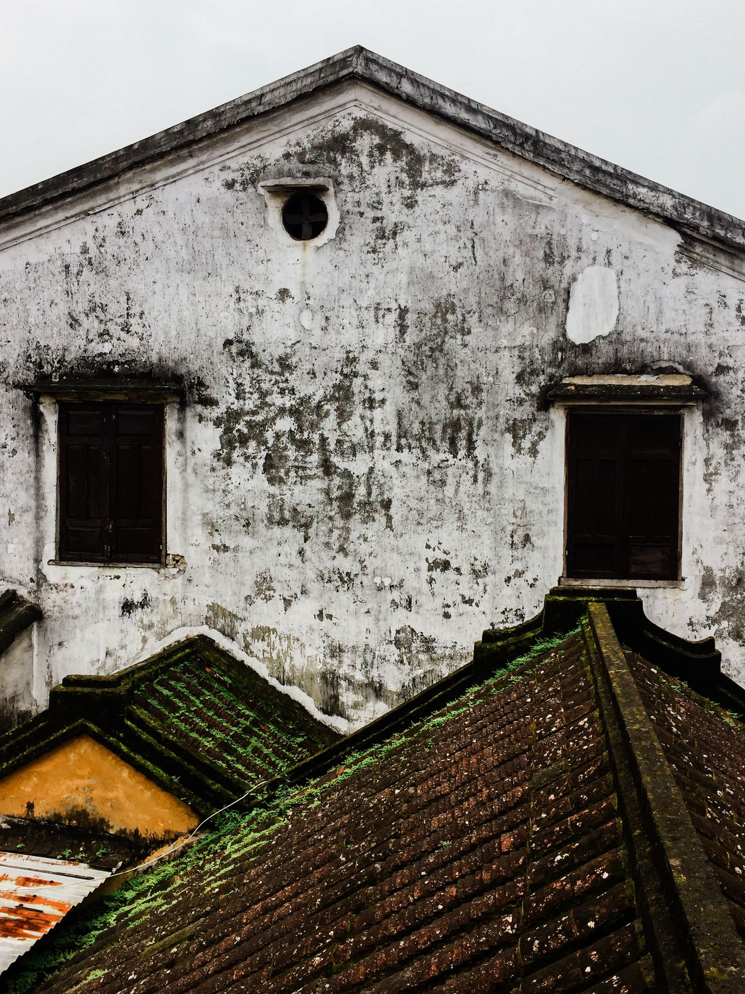 Weathered white building facade with circular window and moss-covered tiled roofs below.