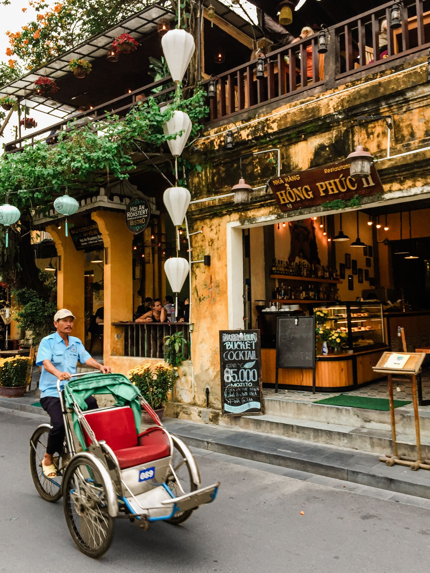 Traditional cyclo rickshaw parked outside a rustic cafe with hanging white lanterns in Hoi An, Vietnam.