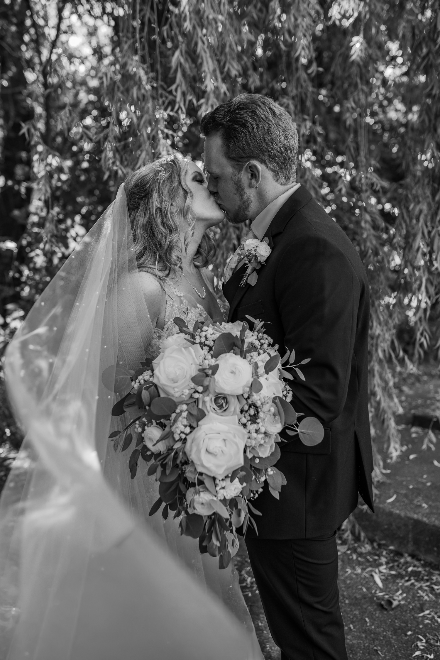 Black and white wedding photo showing a romantic kiss between newlyweds with a beautiful white floral bouquet in a garden setting.