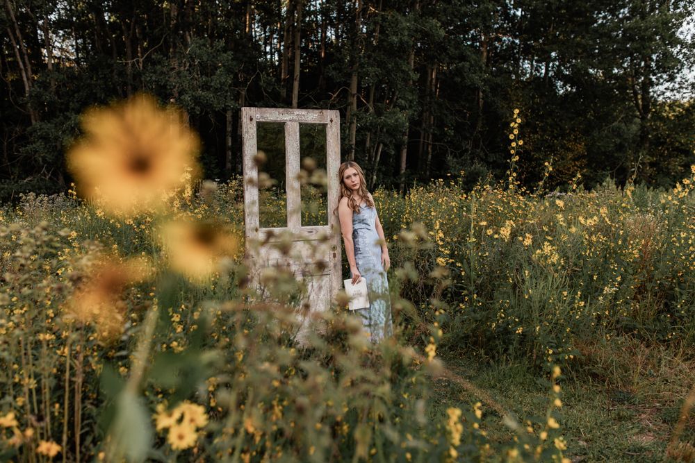 Young woman in floral dress standing by vintage white door frame in wildflower meadow with yellow blooms.