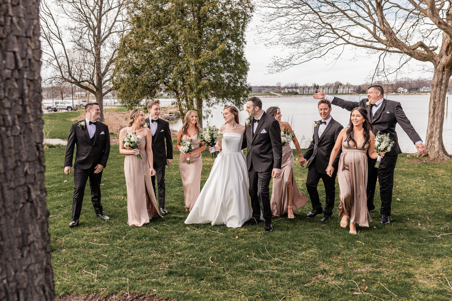 Wedding party walking outdoors near waterfront, bride and groom kissing, bridesmaids in champagne gowns, groomsmen in black tuxedos.